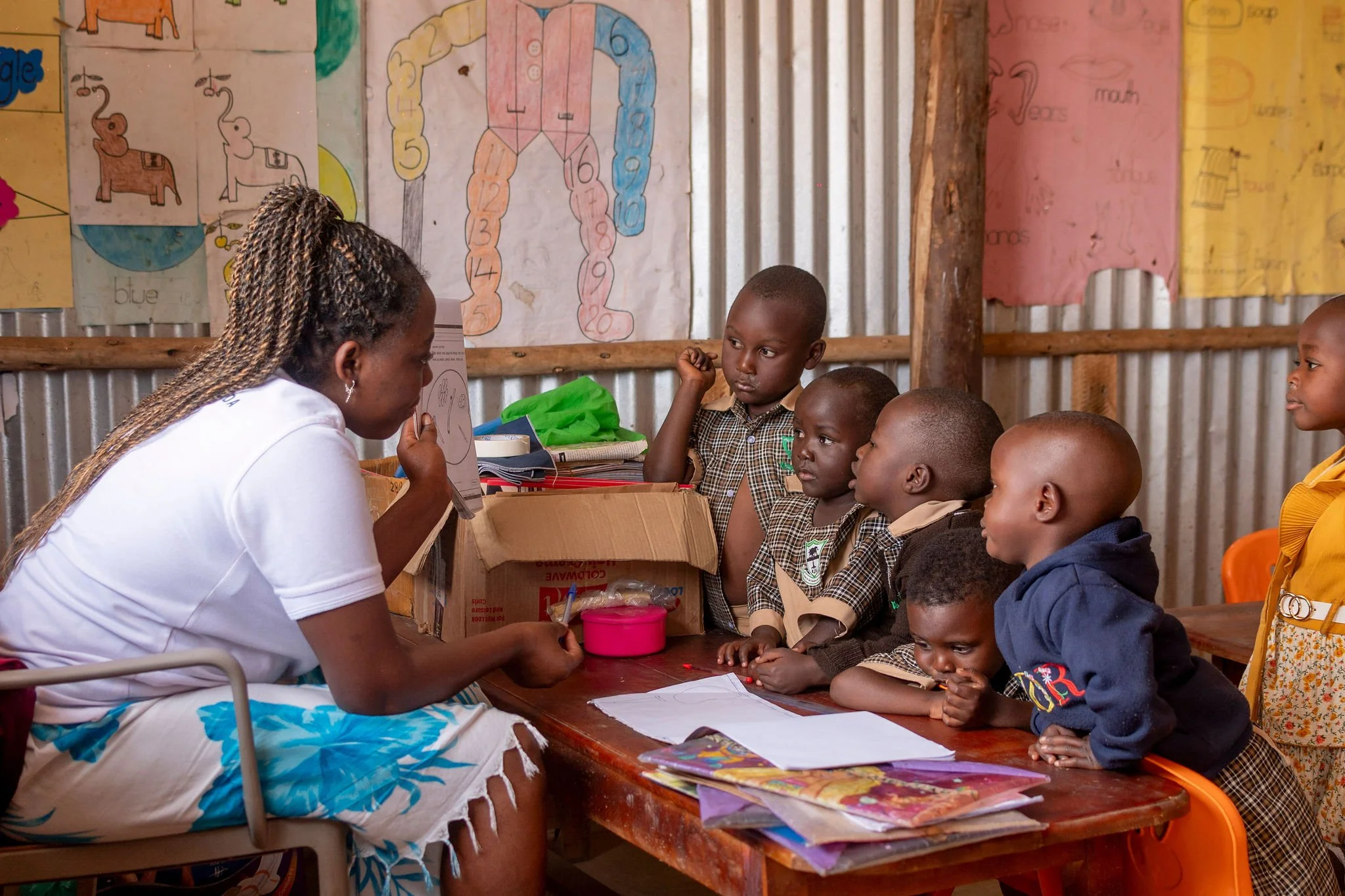 A teacher sitting at a desk with students in a classroom decorated with educational posters about colors and numbers. The students are paying attention to the teacher.