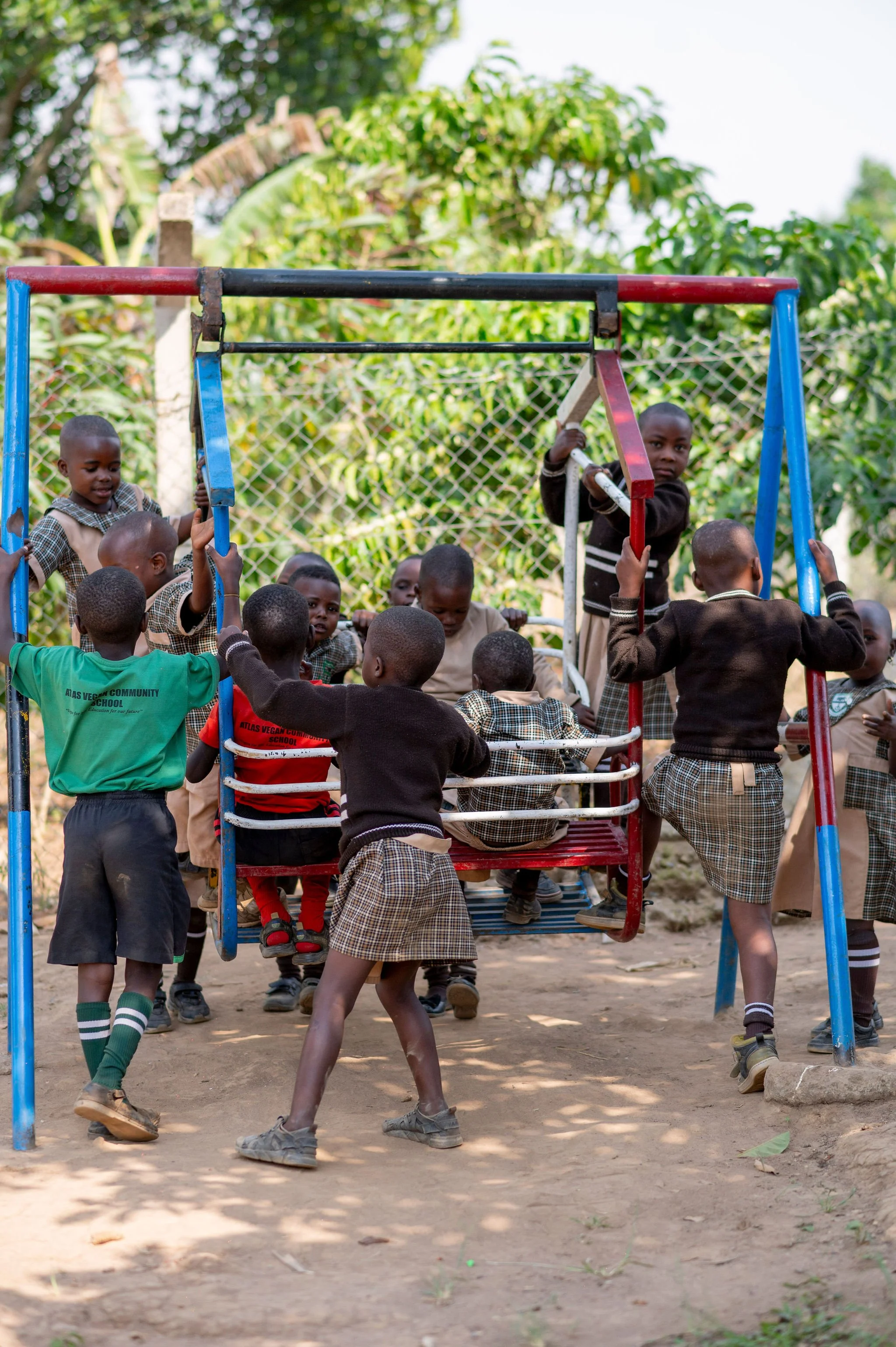 Children playing on a red and blue jungle gym outside on a dirt ground, with trees and a wire fence in the background.