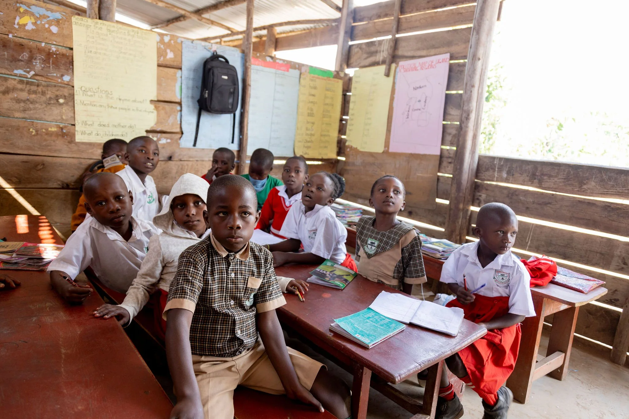 A classroom with about ten children sitting at wooden desks in a wooden-walled room with posters on the wall. The children are looking at the camera, some with serious expressions, and have notebooks and books on their desks.