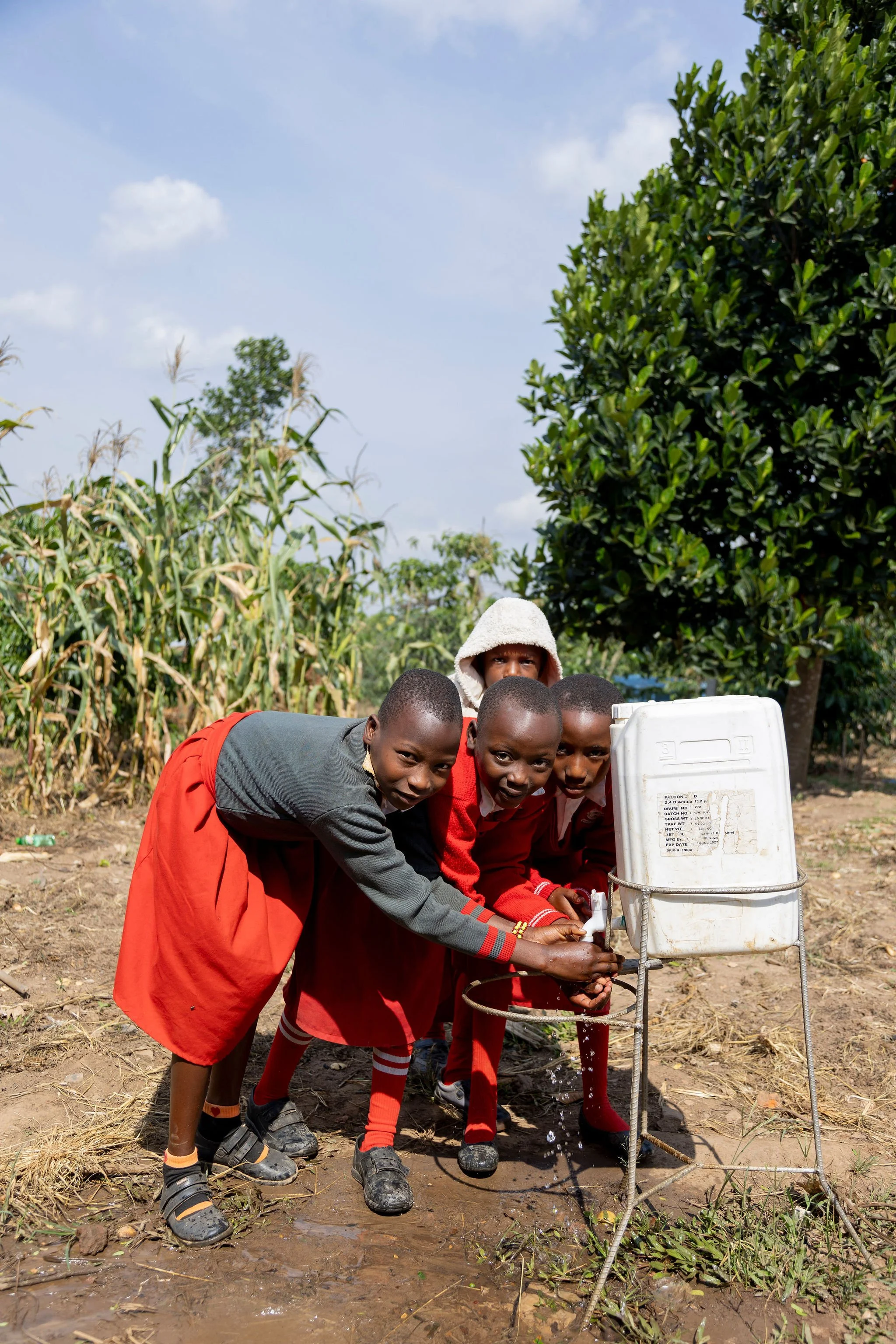 A group of children in school uniforms washing their hands at an outdoor water pump in a rural setting surrounded by trees and crops.