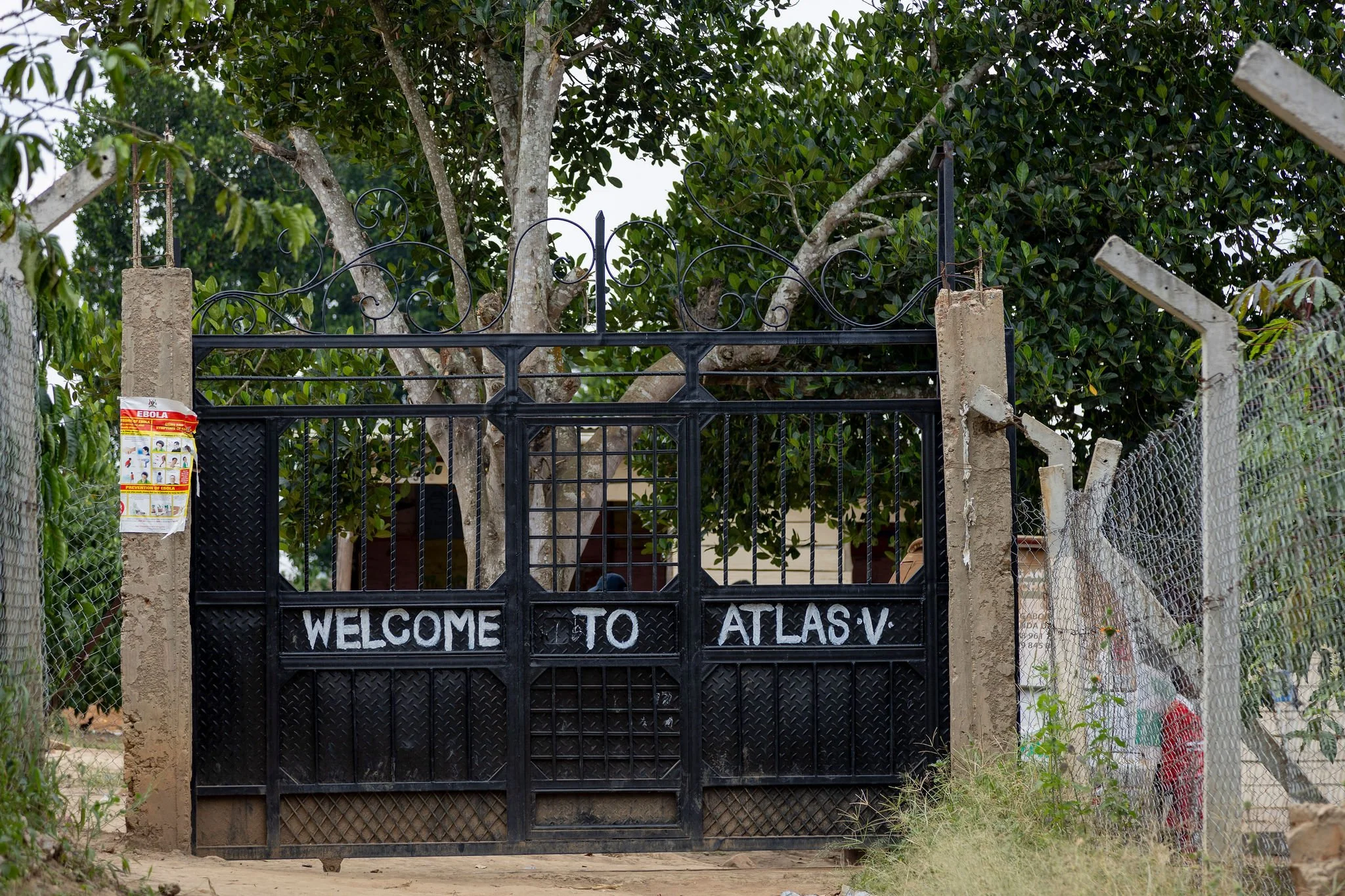 A black metal gate with a welcome sign, flanked by concrete posts, leading to an area with lush green trees and plants. A red and white Ebola prevention poster is attached to one of the posts.