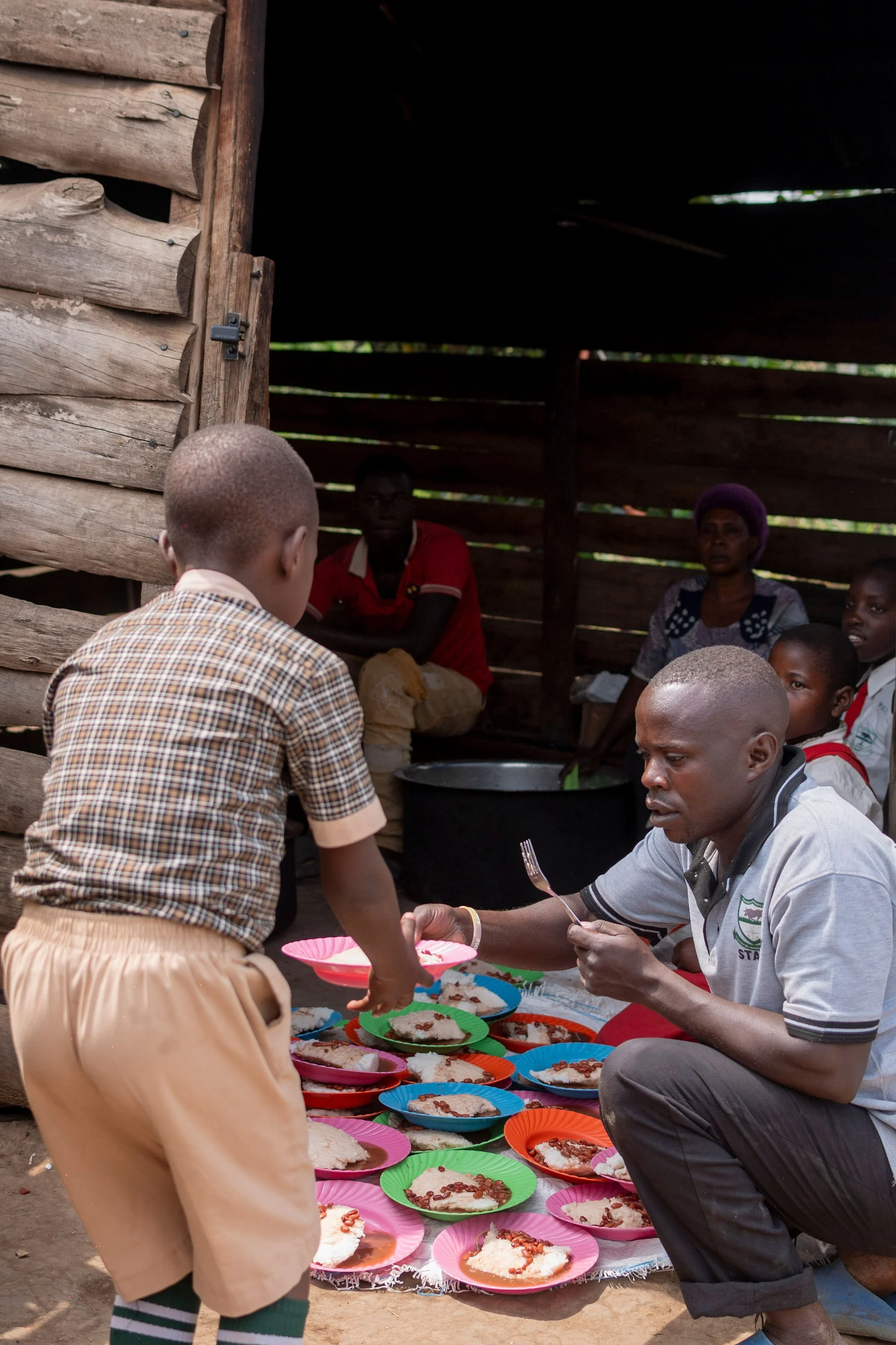 People gathered around colorful plates of food, with a focus on a man handing a plate to a young boy, in a rustic wooden structure with walls made of horizontal planks.