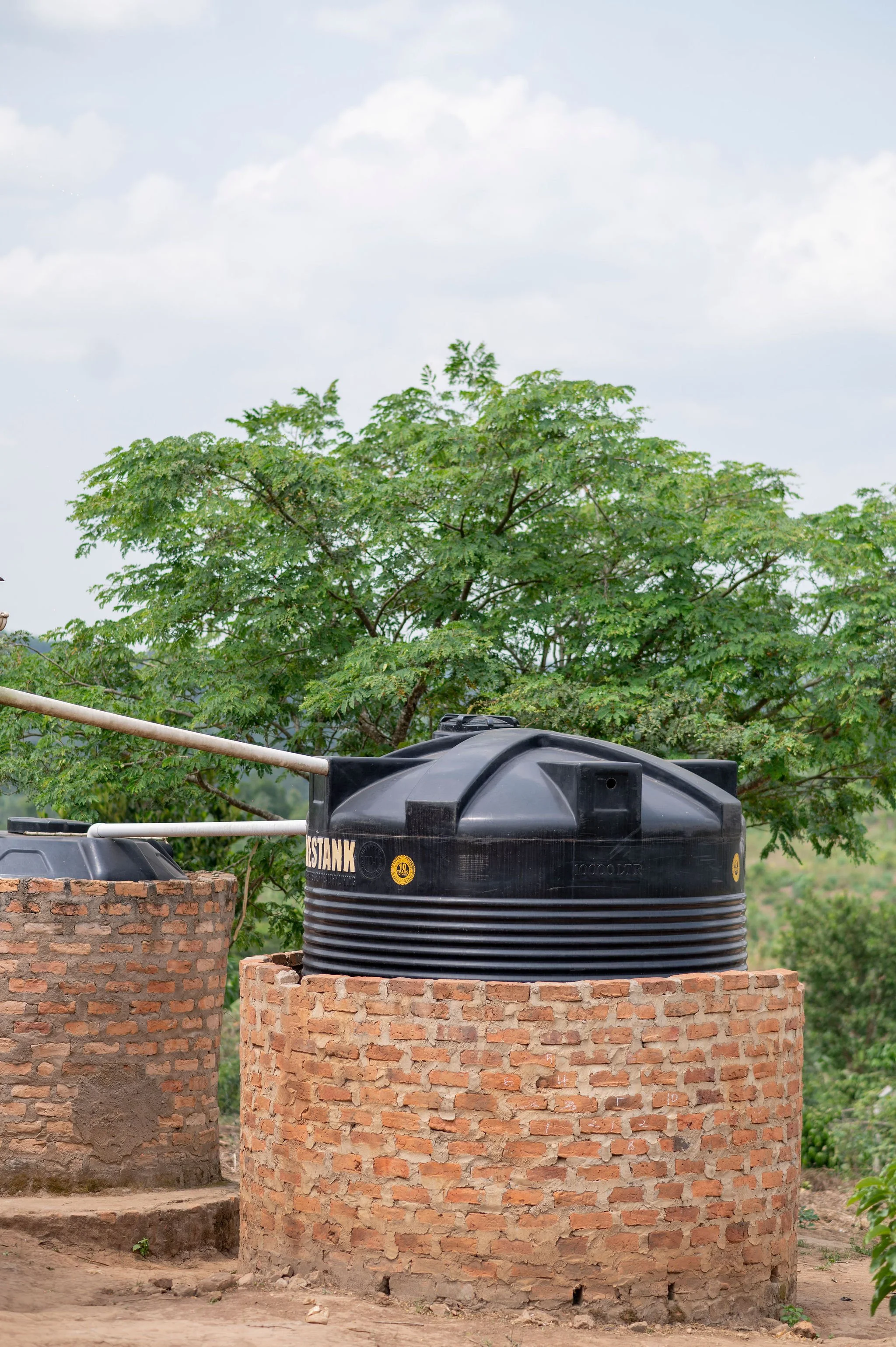 Two brick water tanks on a dirt ground, with one black water tank placed on a surrounding brick wall structure. There is lush green foliage and trees in the background, with a cloudy sky overhead.