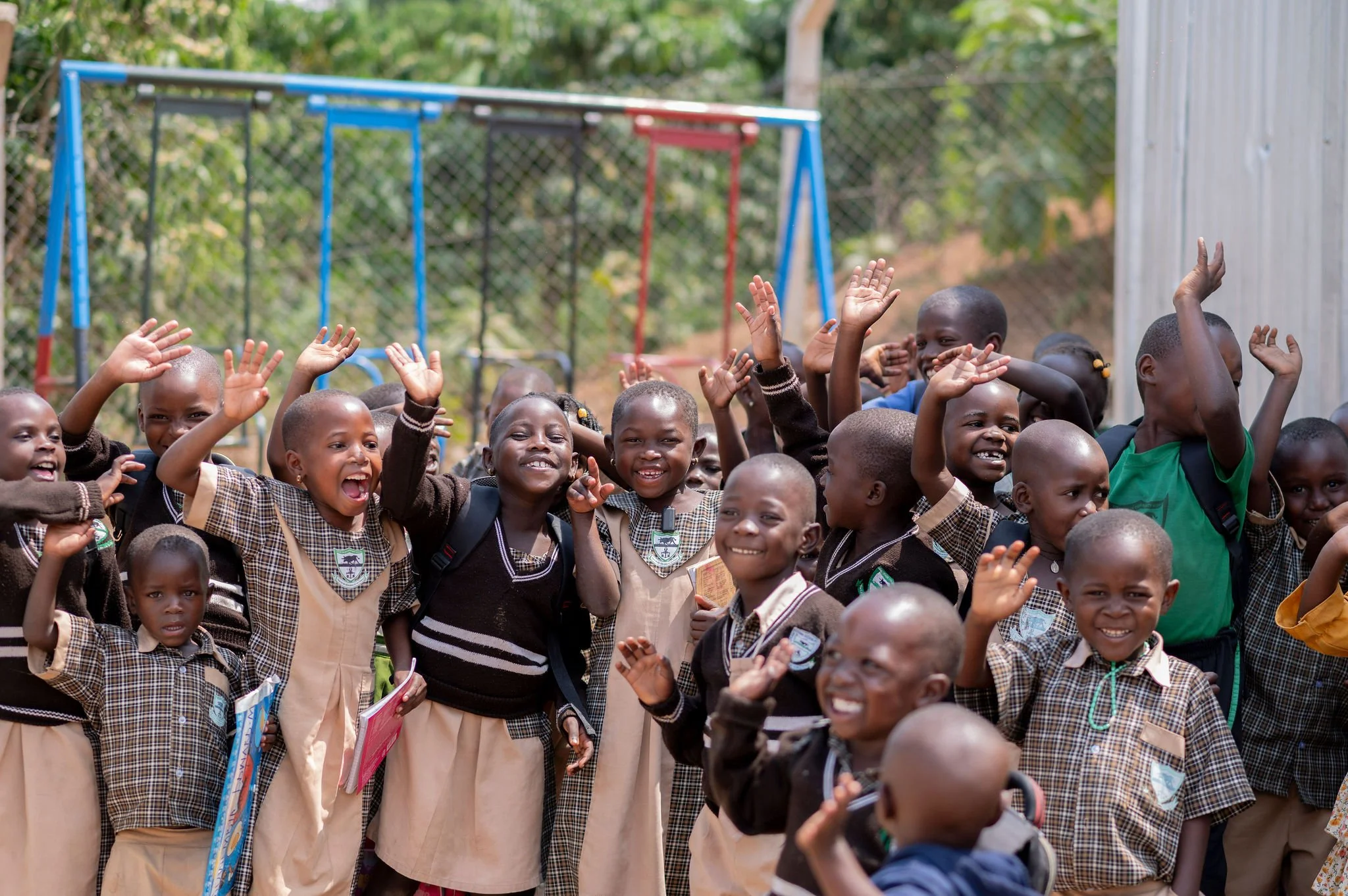 Group of smiling children in school uniforms waving and raising their hands outdoors near a playground.