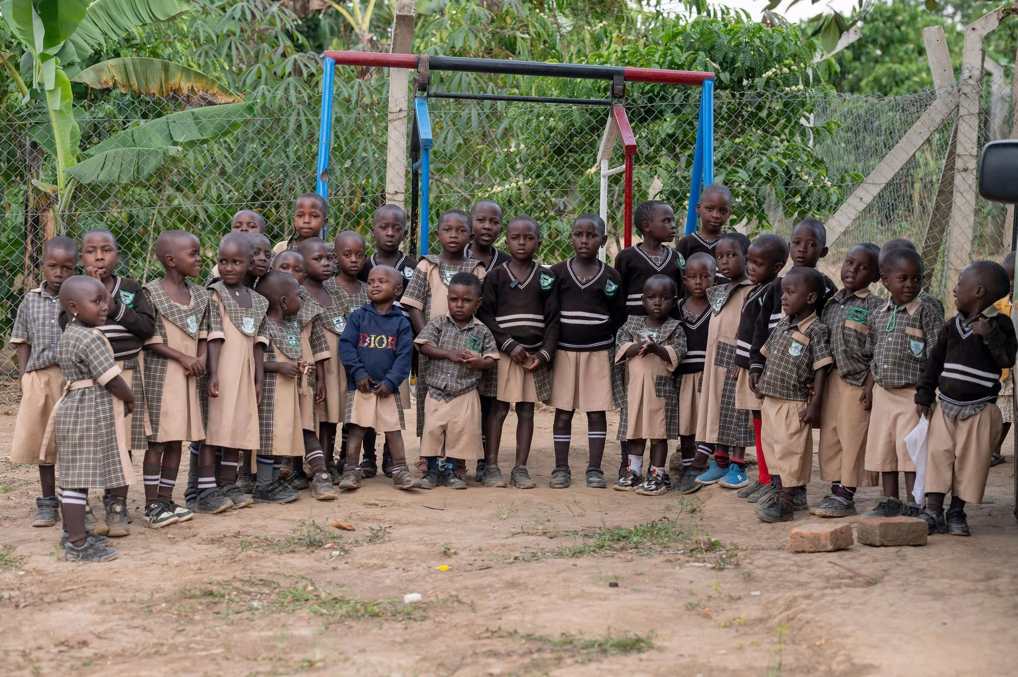 Group of children in school uniforms standing outdoors near a chain-link fence and a jungle-like background.