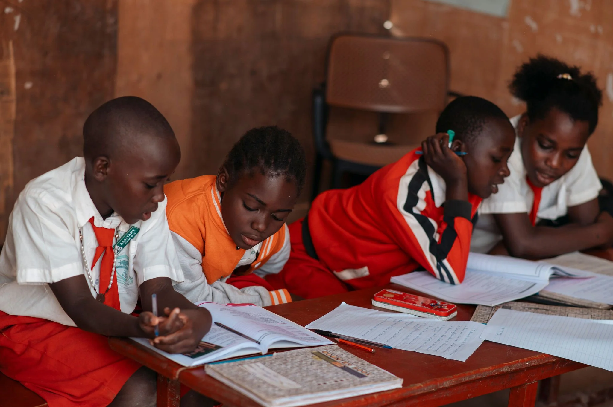 Group of students sitting at a desk, studying with books, papers, and writing utensils, in a classroom with a wooden wall.