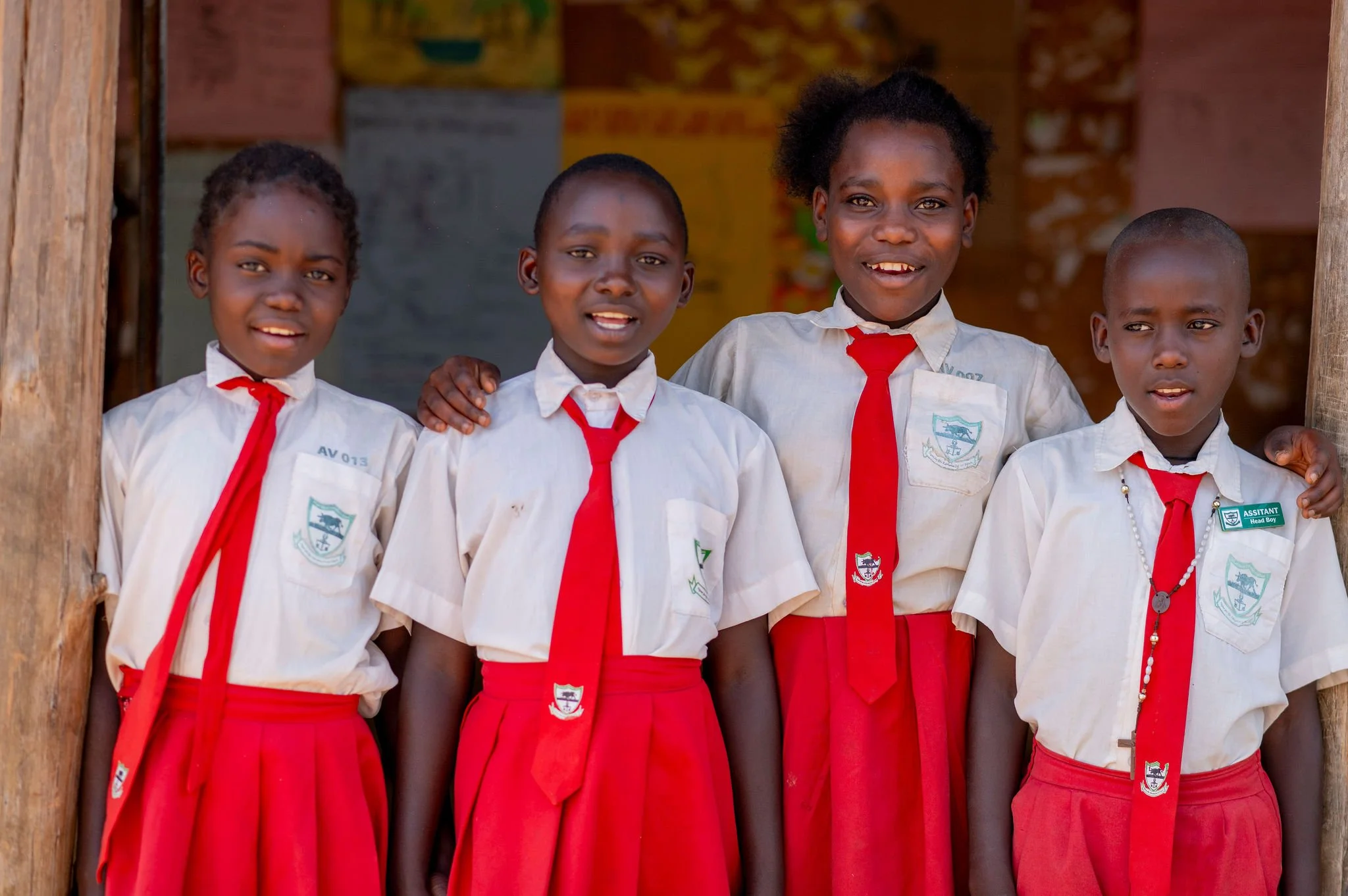 Four children in school uniforms, standing together and smiling, outside a wooden structure.