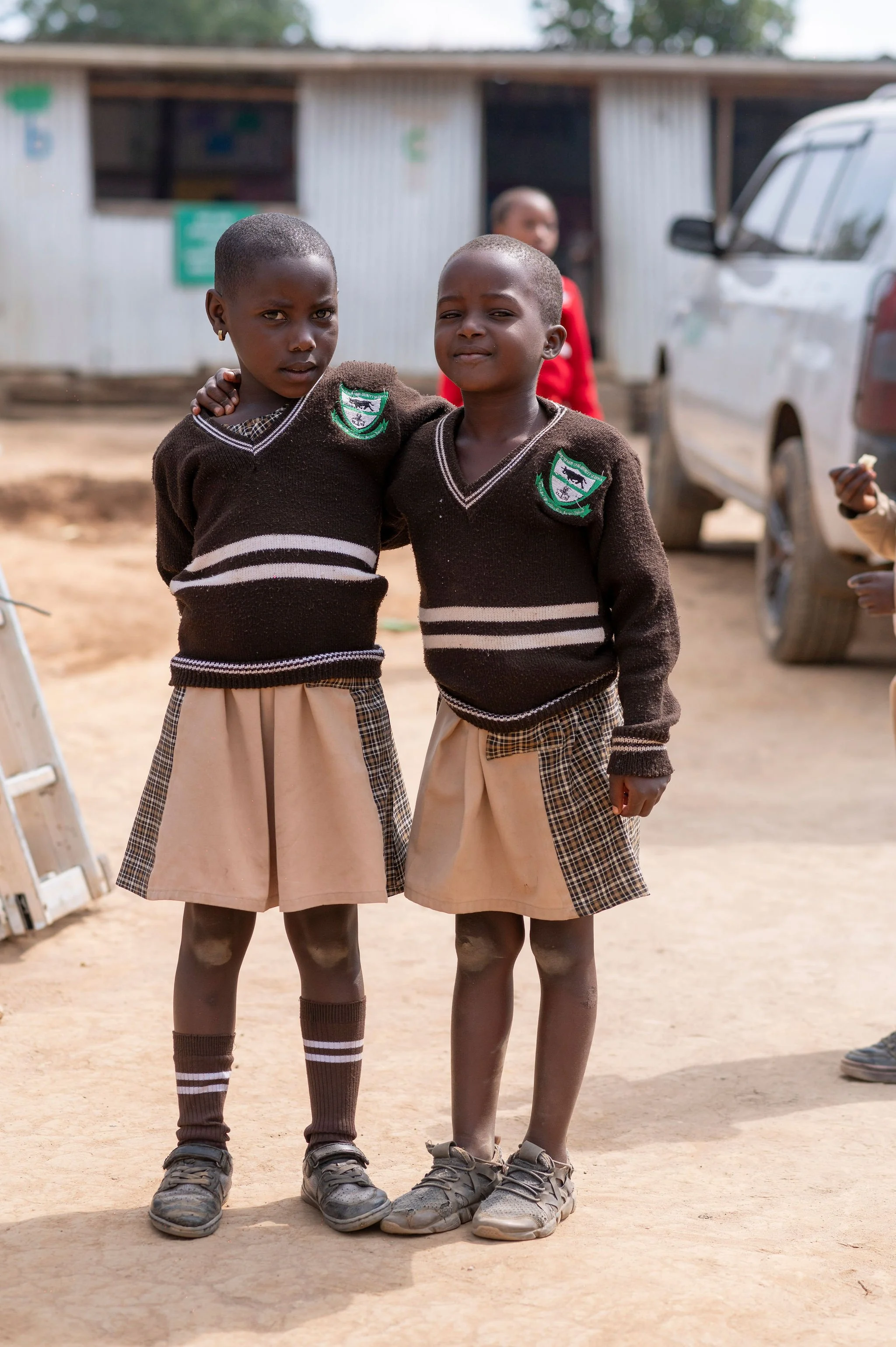 Two young schoolgirls in matching uniforms standing side by side outdoors, embracing each other, with a building and vehicles in the background.