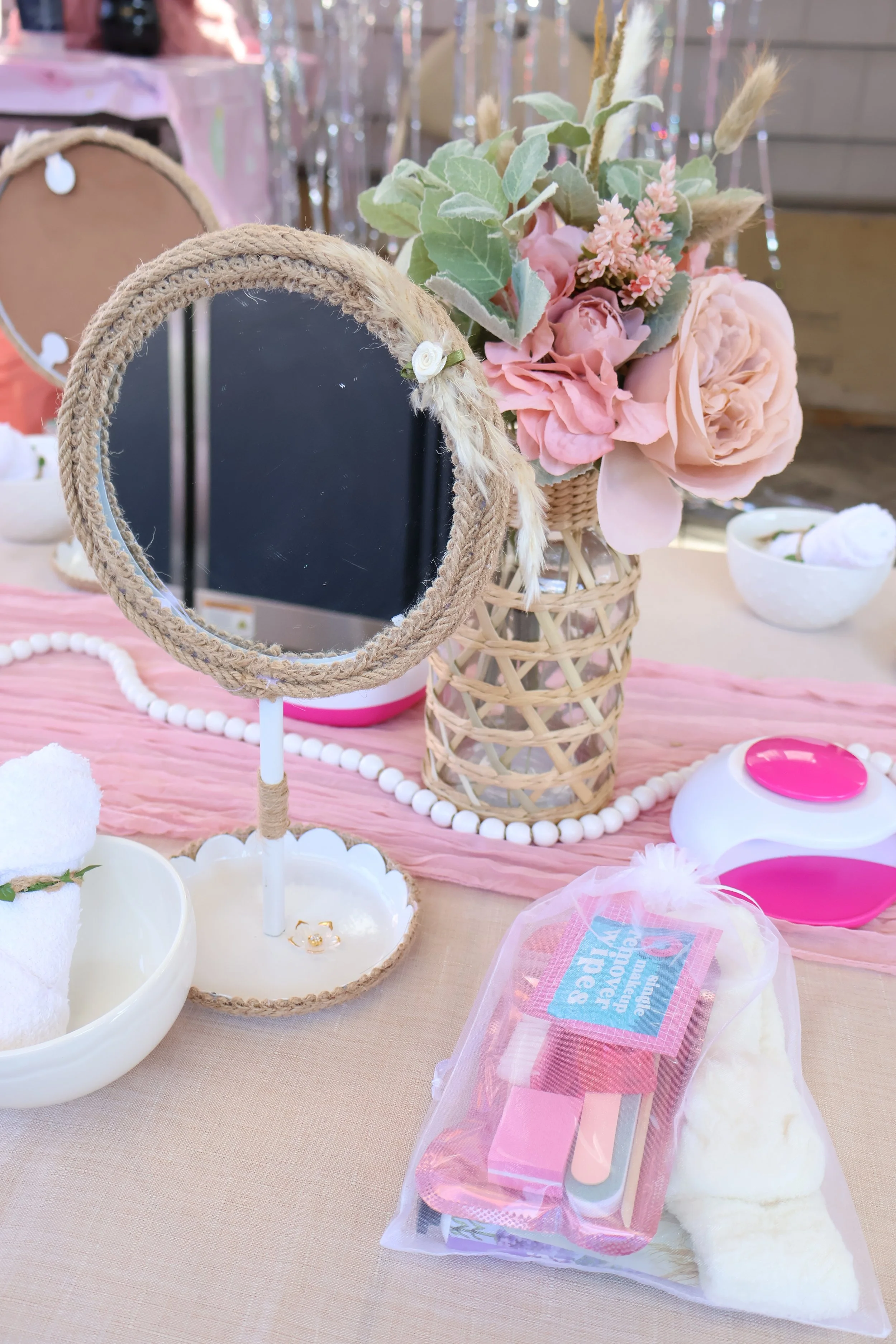 A pink-themed table setting with a bouquet of pink flowers and green leaves, a small mirror wrapped in twine, and various bath and beauty items including a loofah, bath bombs, and a container of cotton pads.