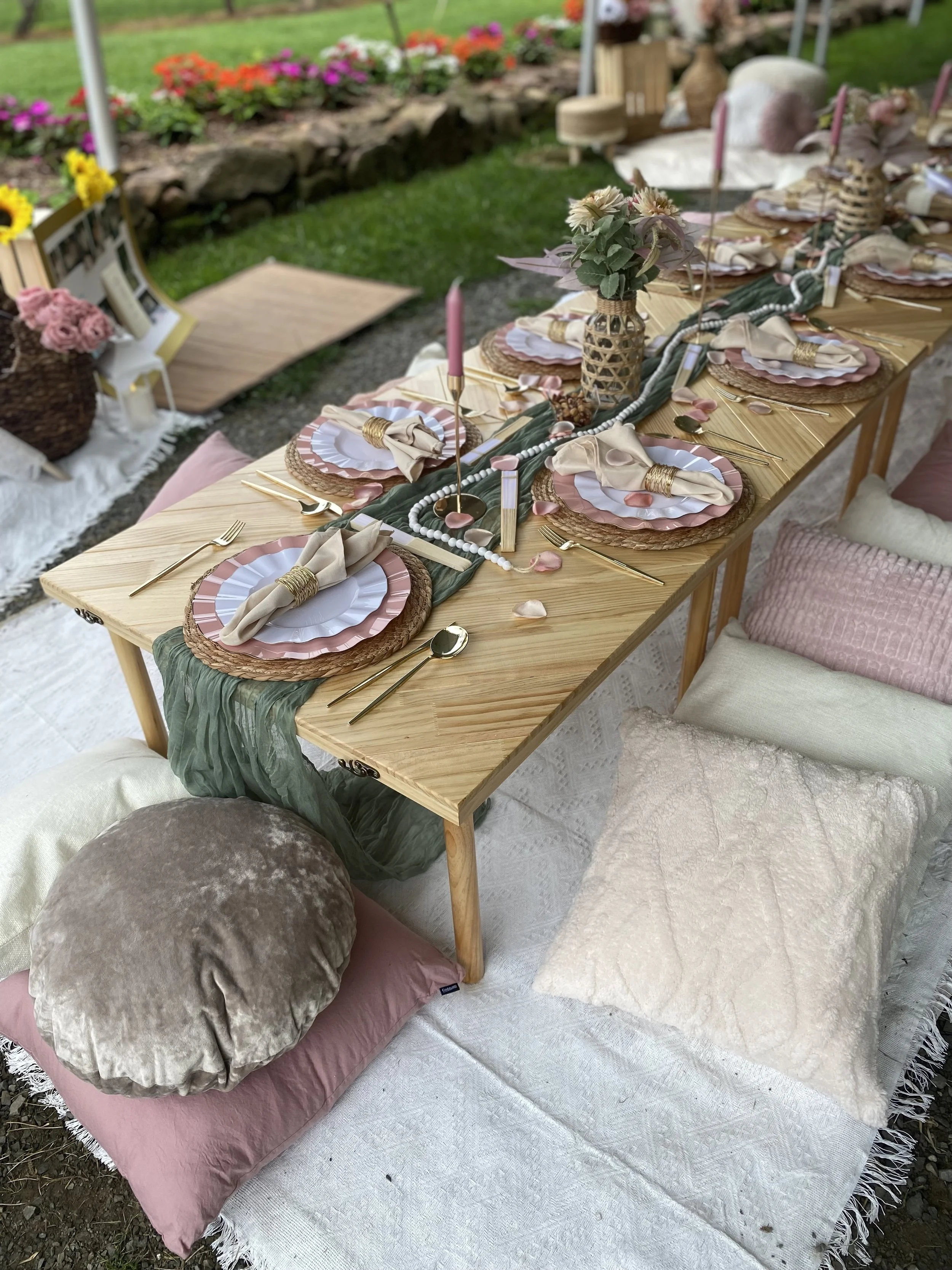 A decorated outdoor dining table set with pink, beige, and green tableware, candles, and floral arrangements, accompanied by plush cushions and pillows for seating, with a garden flower border in the background.
