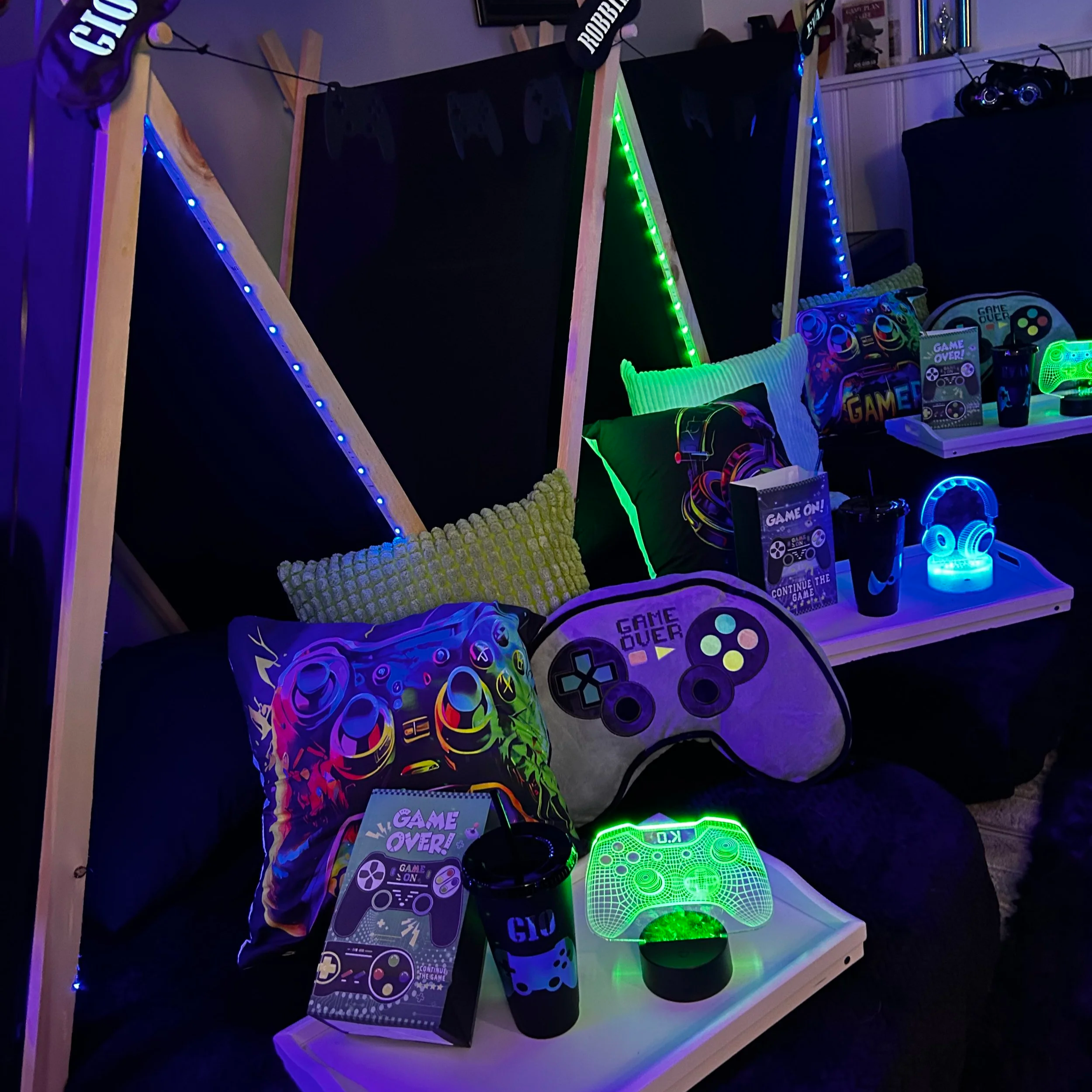 Colorful gaming-themed decorations and LED lights on a shelf, including game controllers, pillows, and signs with video game graphics, in a room with purple and blue lighting.
