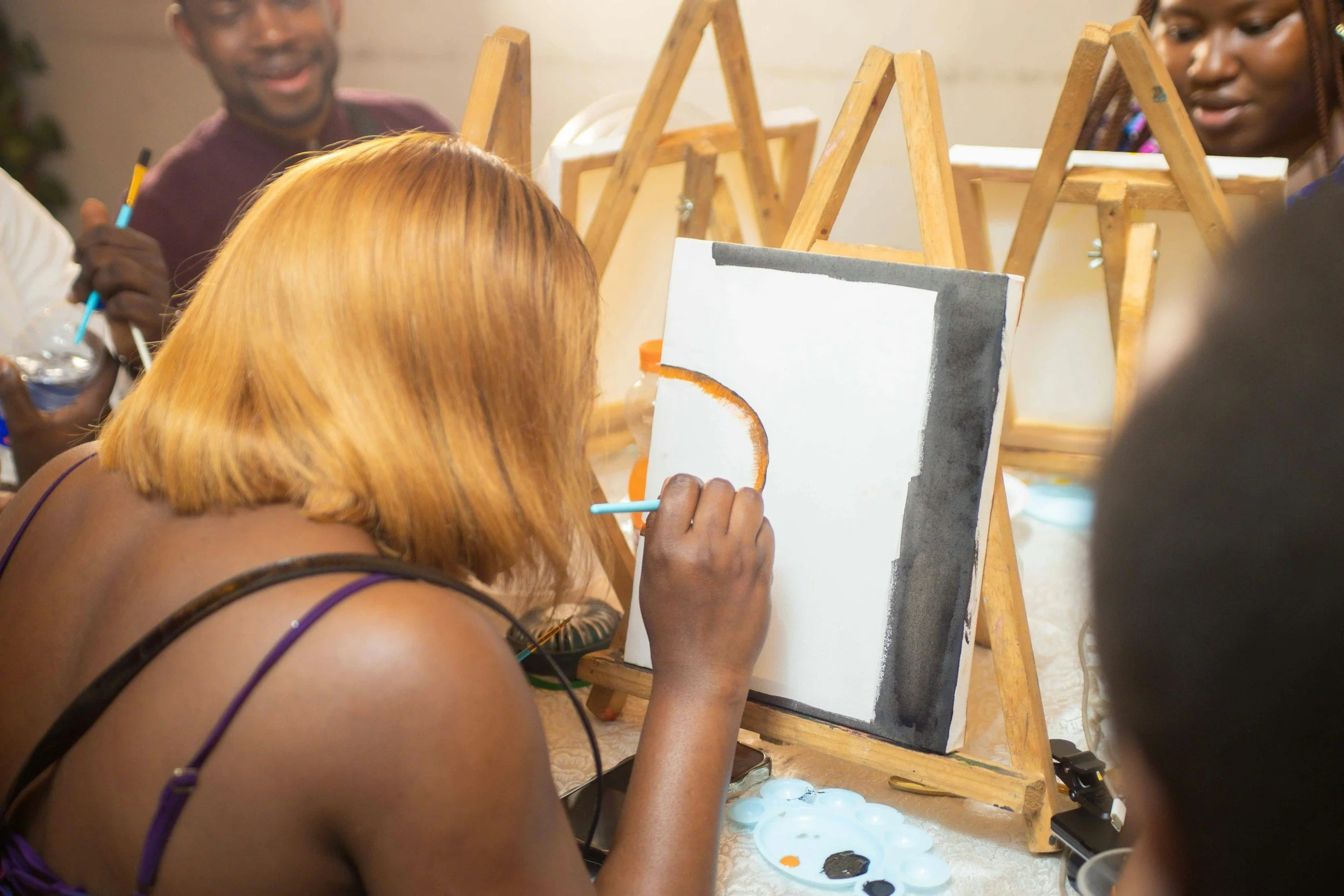 A woman with blonde hair painting on a canvas at an art class, with other people in the background and easels around.