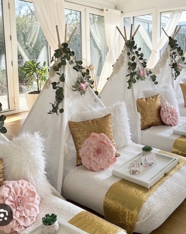 Cozy indoor space decorated with teepees, gold pillows, fluffy and floral pillows, and greenery, near large windows with white curtains.