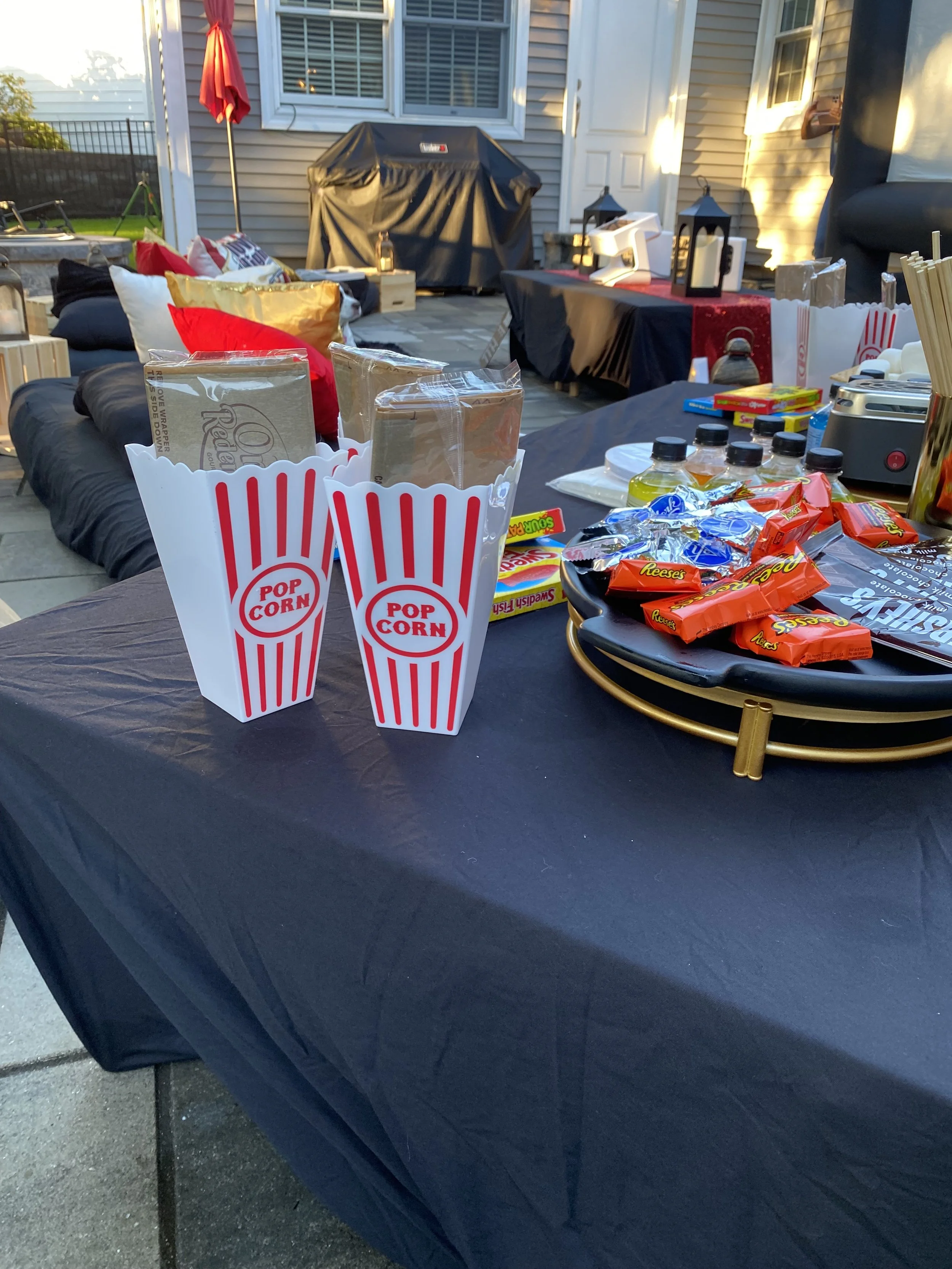 Outdoor scene of a backyard patio set up for a gathering with snacks and decorations. Popcorn and candy bags on the table, pillows on the seating area, and a grill in the background.