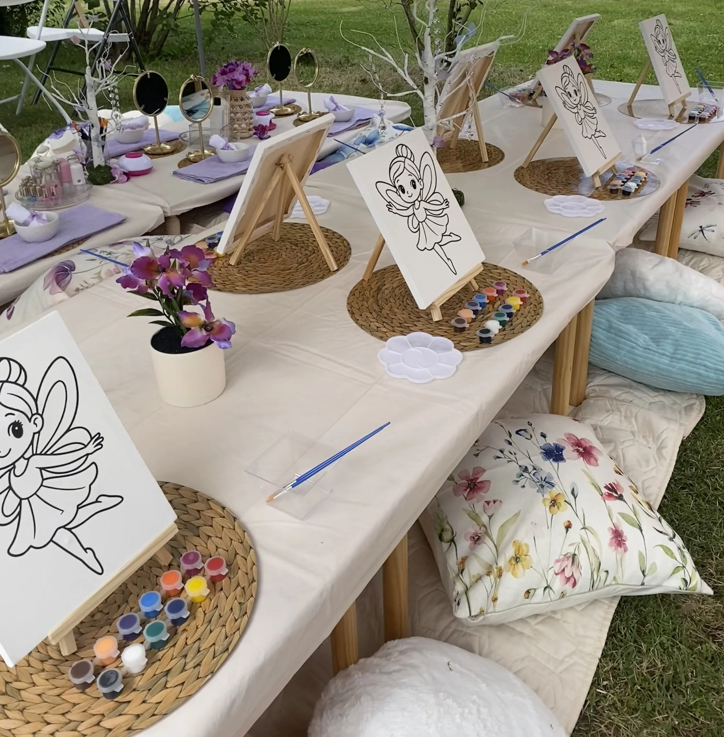 Outdoor activity table with painting supplies, watercolor paints, small canvases, mirrors, and purple flowers, set on a white tablecloth on grass.