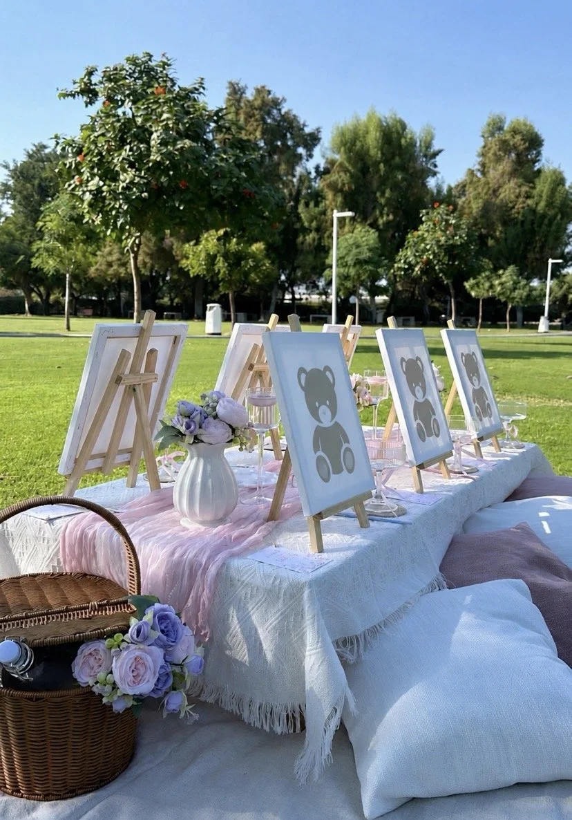 Outdoor setup with small canvas paintings of teddy bears on easels, floral arrangements, and picnic blankets on grass.