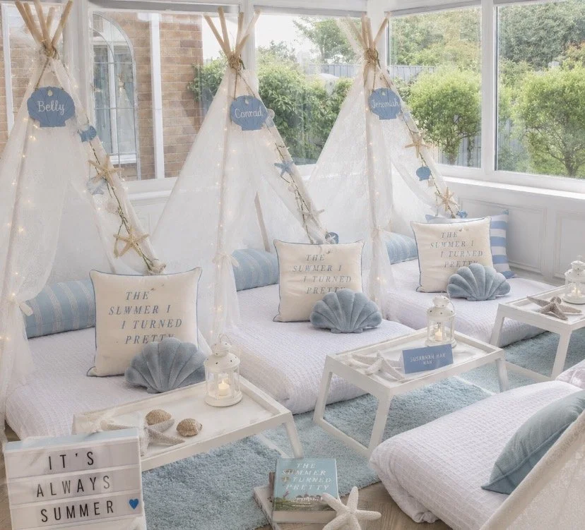Indoor summer-themed lounge area with white tents decorated with starfish and string lights, each labeled with names 'Belly,' 'Carmel,' and 'Jeremiah.' Cushions and seashell decor are on the floor and small white tables in front of the tents, creating a beach-inspired atmosphere.
