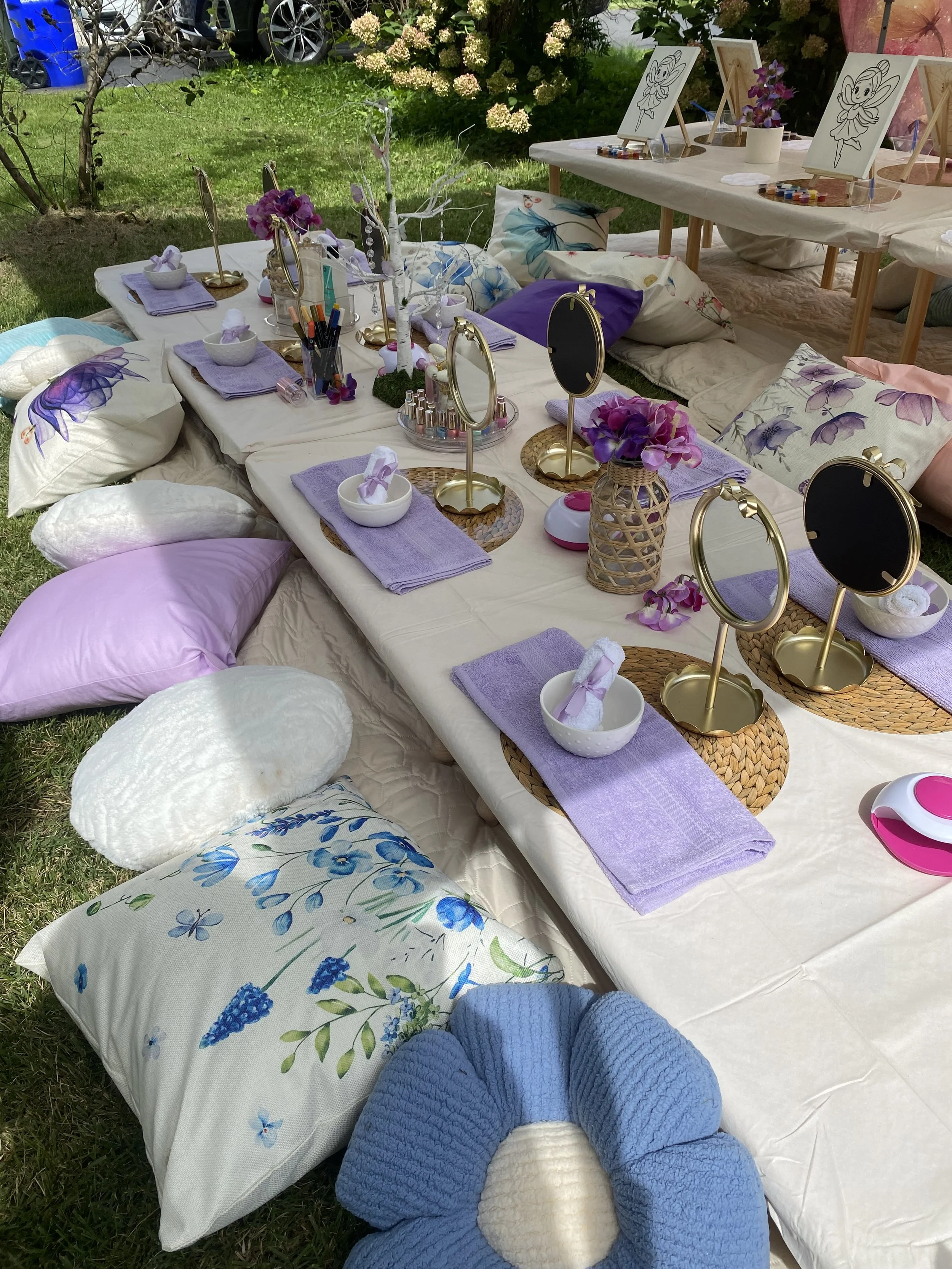 An outdoor craft or tea party setup with pillows, small tables, and art supplies on a grassy area with trees and cars in the background.