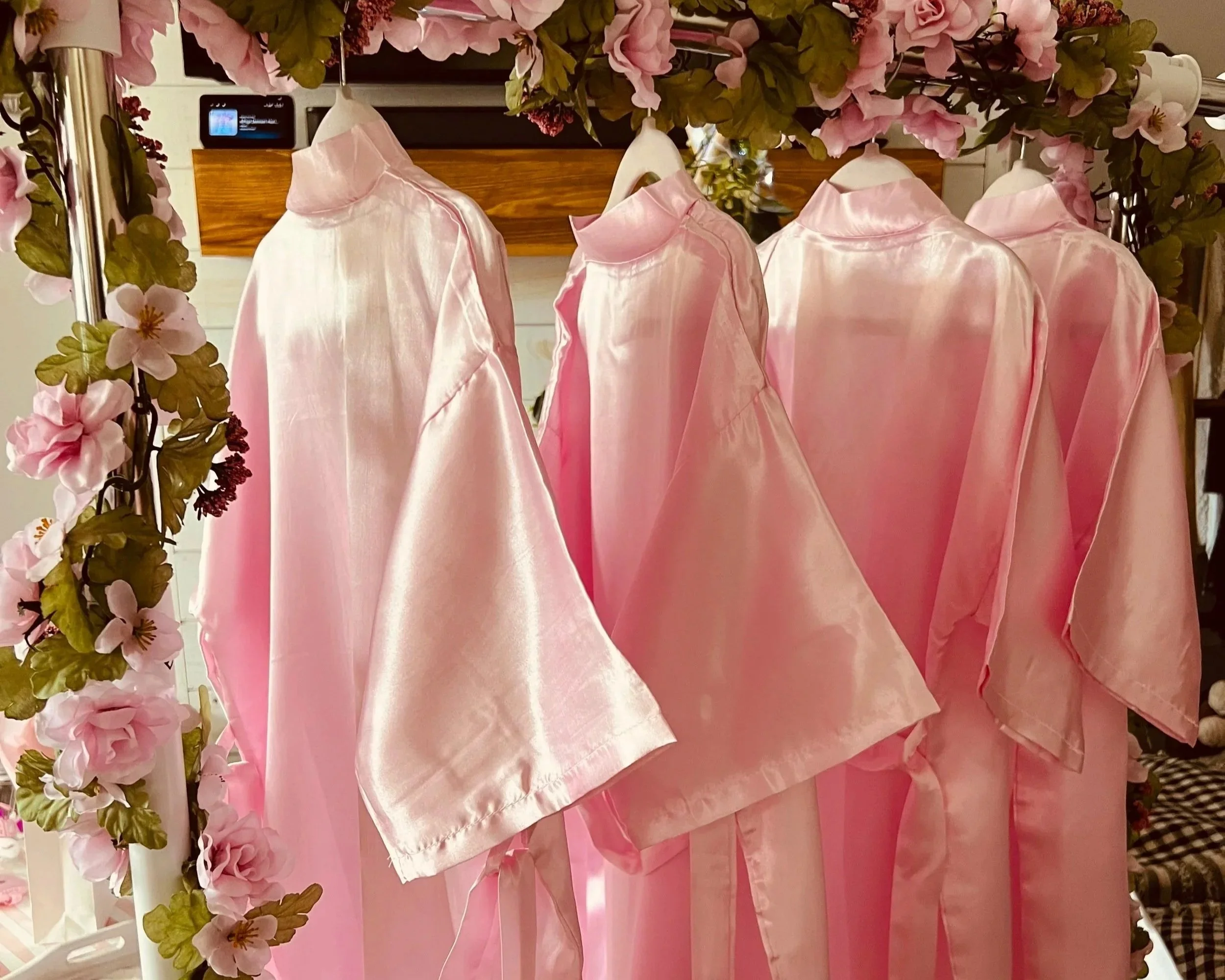Pink satin shirts hanging on a floral clothing rack, with pink and green flowers on the sides.