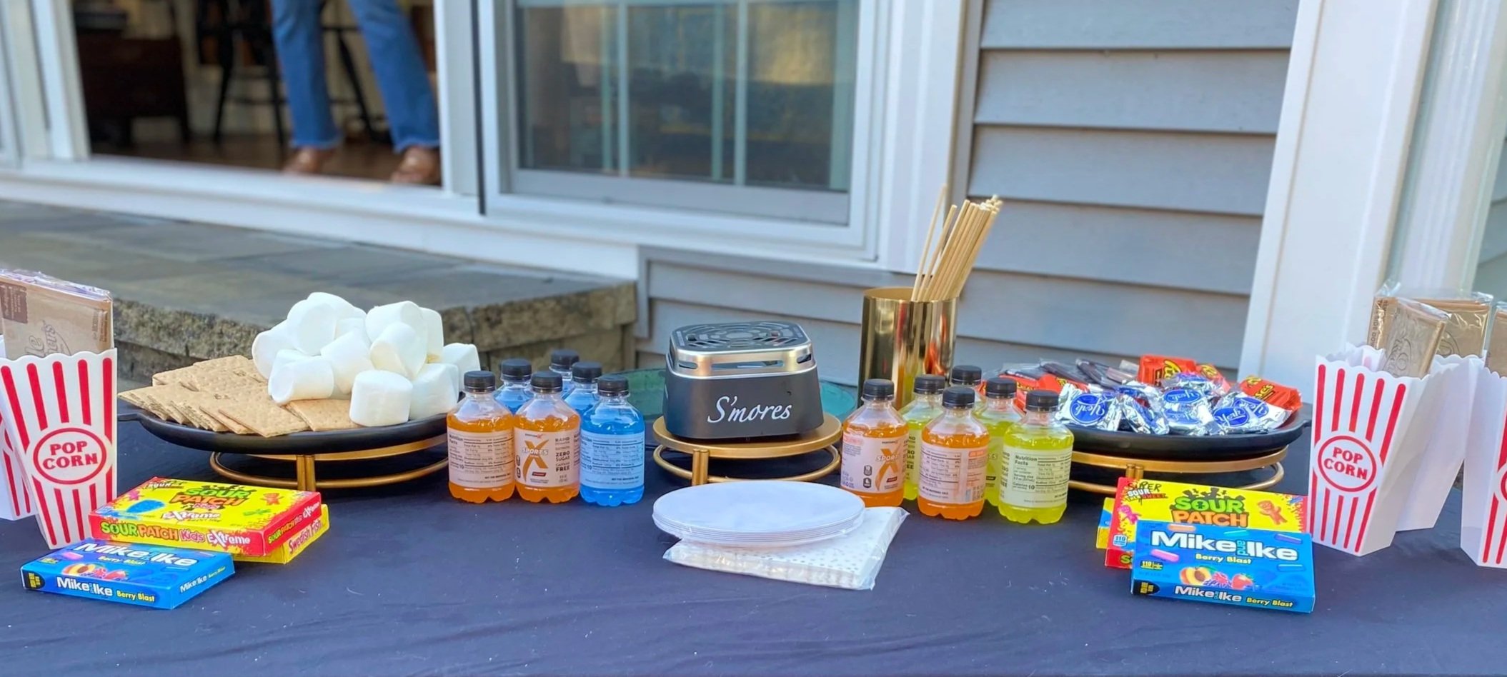 Outdoor table with popcorn, s'mores supplies, colorful drinks, and candies for a snack station.