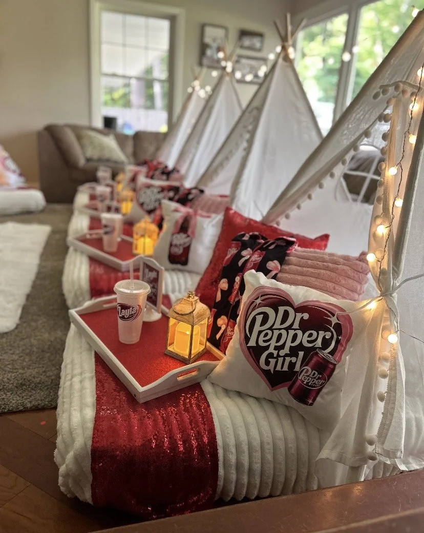 A cozy indoor setting featuring a row of white teepees decorated with string lights, each with a pillow and tray containing a Lay's drink and a lantern. A red and white striped blanket drapes over the beige couch in the background.