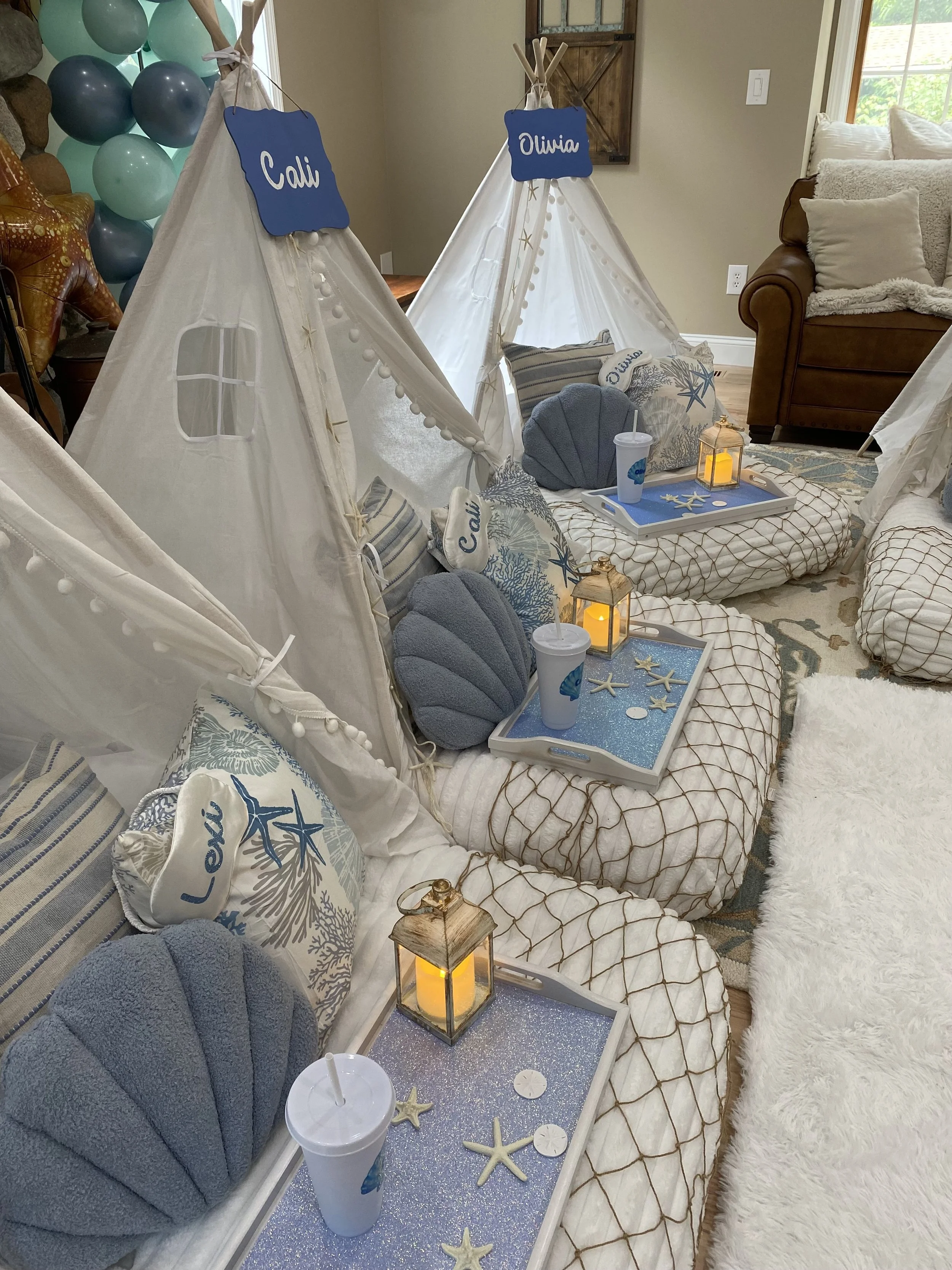 Beach-themed indoor setup with two white canvas tents labeled 'Cali' and 'Olivia', decorated with starfish and sea-themed pillows, surrounded by blue and white accents, lanterns, and drinks with straws, on a sparkly blue table.