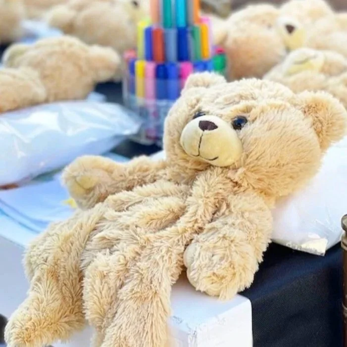 A beige teddy bear lying on a table with other teddy bears and a container of colorful markers in the background.