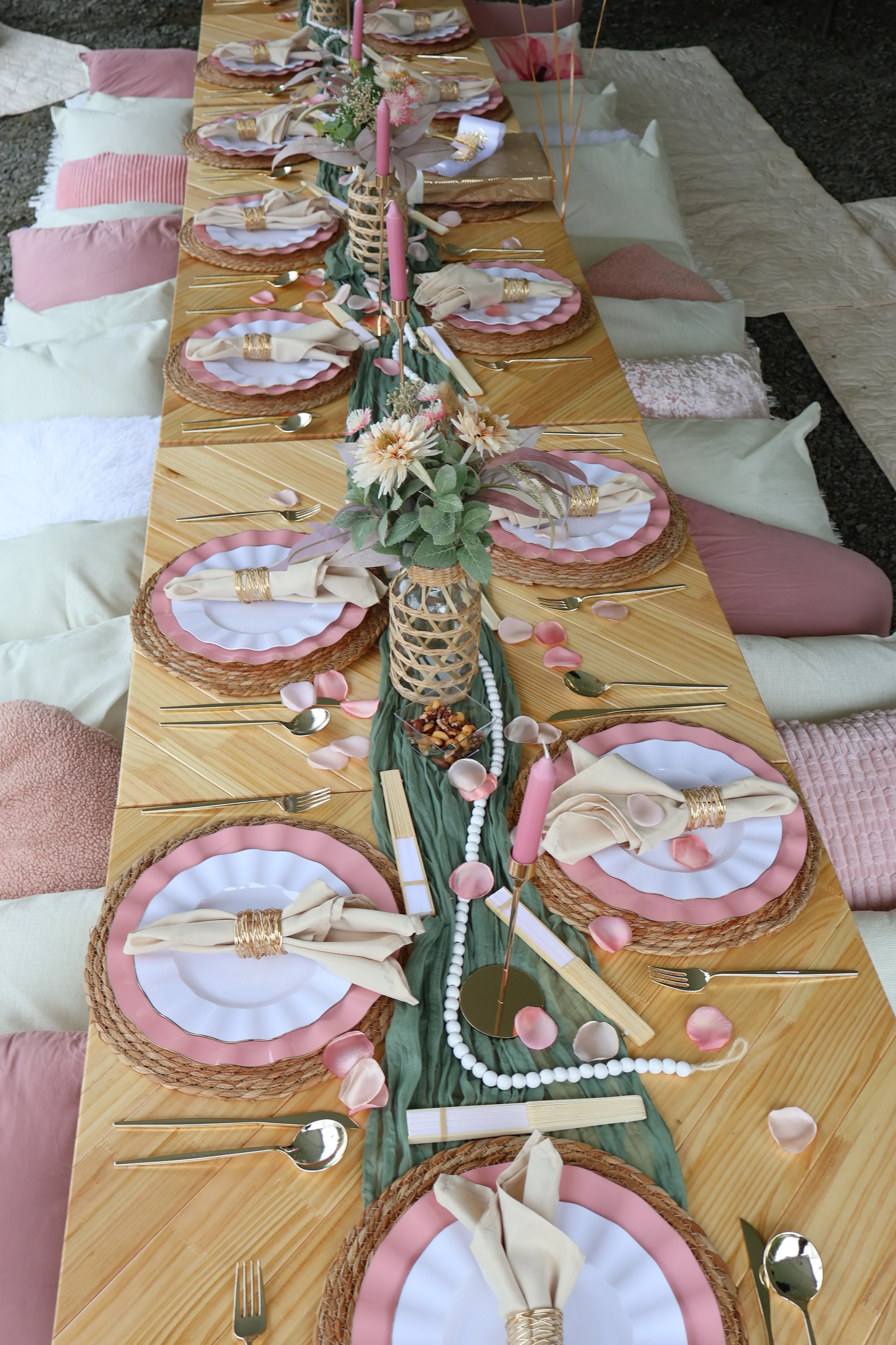 Table setting with pink, white, and gold accents, including plates, napkins, cutlery, candles, and floral decorations, surrounded by floor seating with cushions.