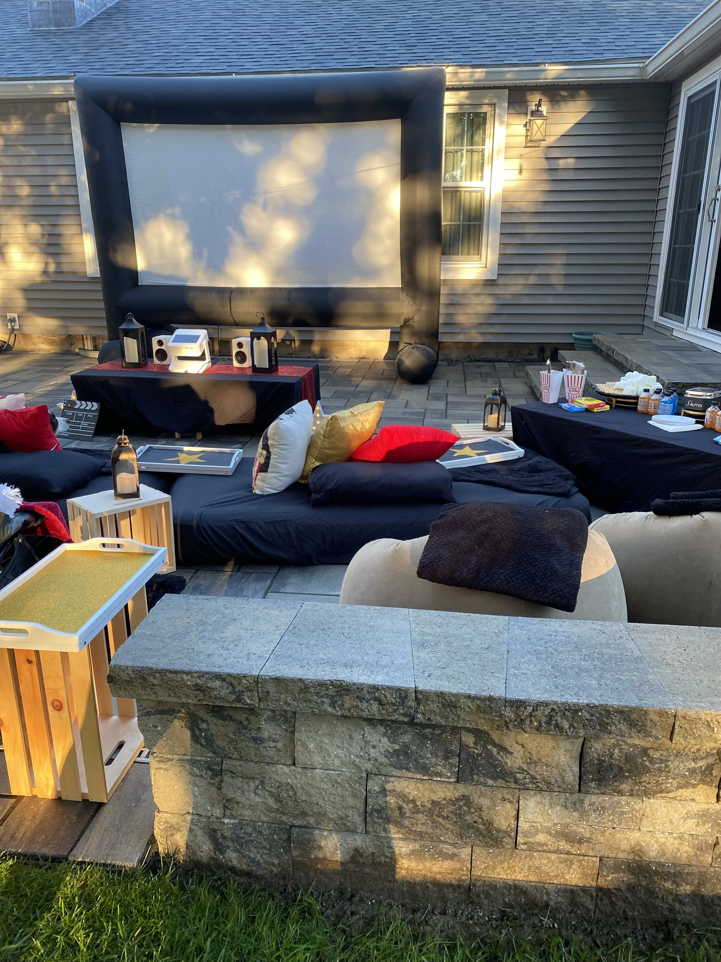 Backyard setup with large inflatable outdoor movie screen, seating area with cushions and pillows, two tables with lanterns, snacks and drinks, and a stone wall in the foreground. The scene is lit by outdoor lighting, indicating evening time.