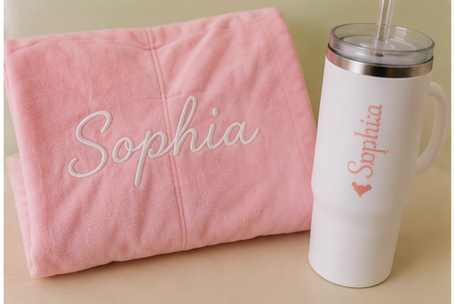 A pink blanket with the name 'Sophia' embroidered on it and a white insulated tumbler with 'Sophia' and a heart design, with a straw.
