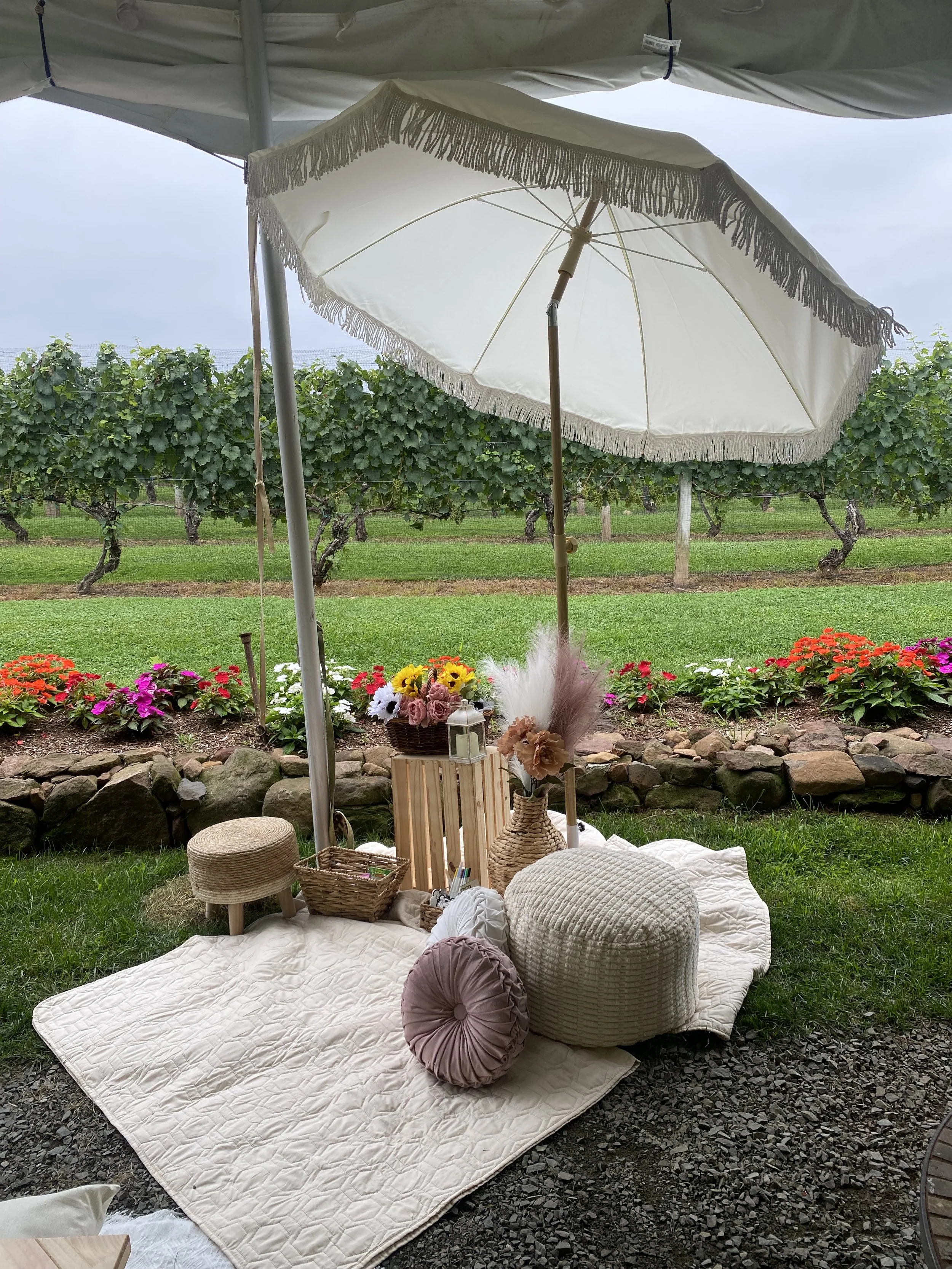 A cozy outdoor picnic area with a white quilted blanket, pillows, woven baskets, a small stool, and decorative flowers, shaded by a large white umbrella on a grassy lawn with a vineyard and cloudy sky in the background.