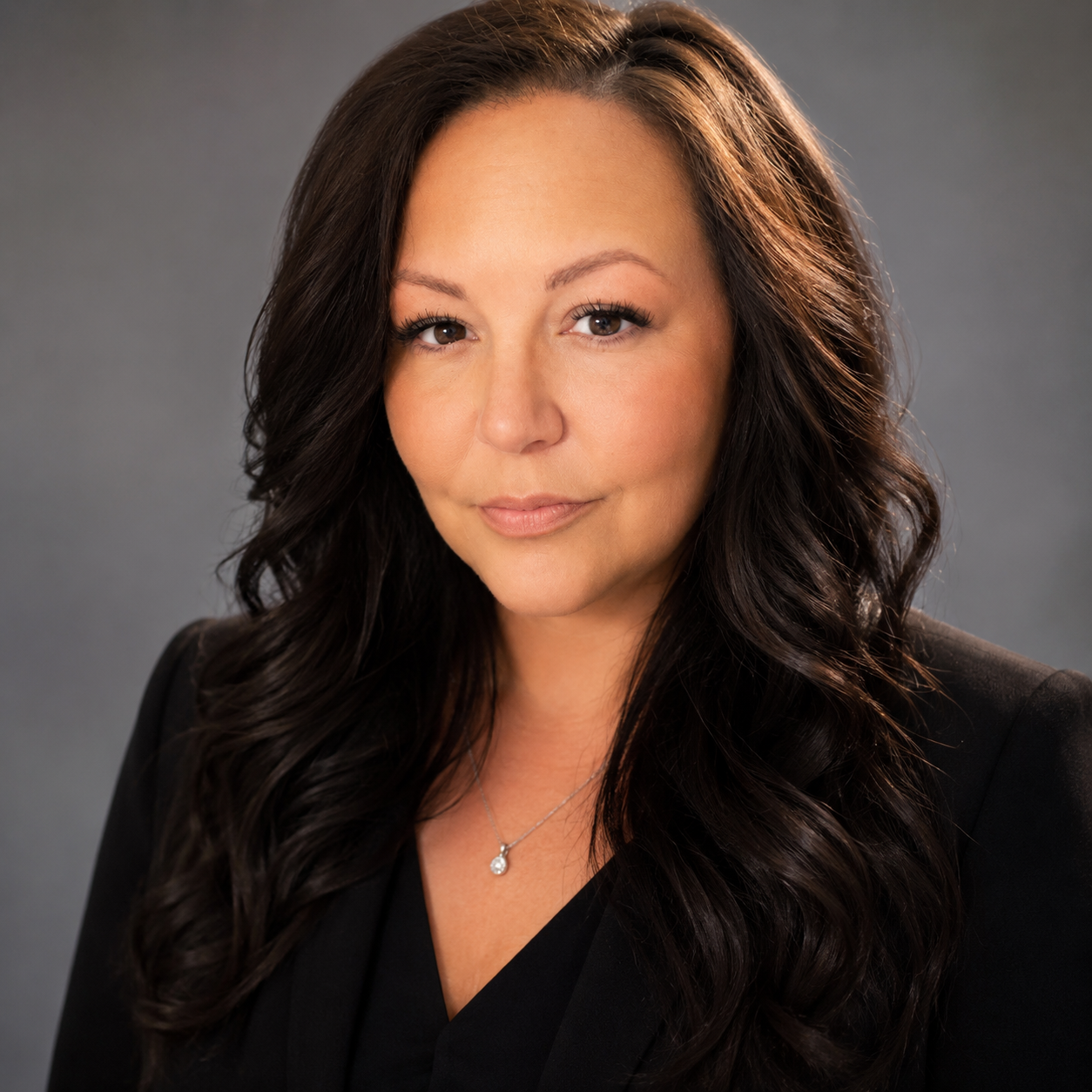 Portrait of a woman with long dark hair wearing a black blazer and a silver necklace, looking confidently at the camera against a gray background.