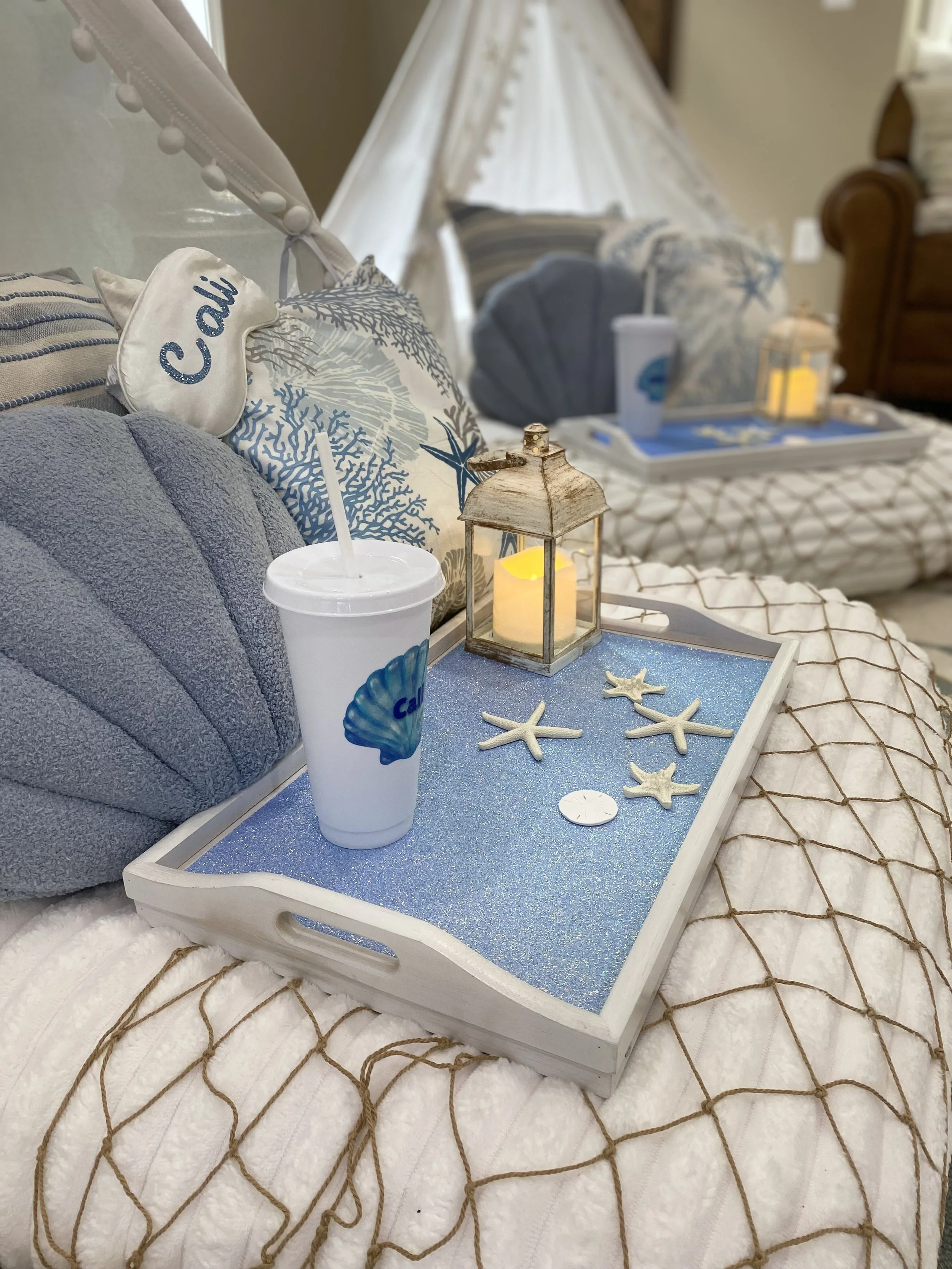 A bedroom decorated with a beach theme, featuring pillows with marine designs, a lantern with a candle, starfish decorations, and a tray with a drink and a lamp. The bed has a white quilt with a brown diamond pattern.