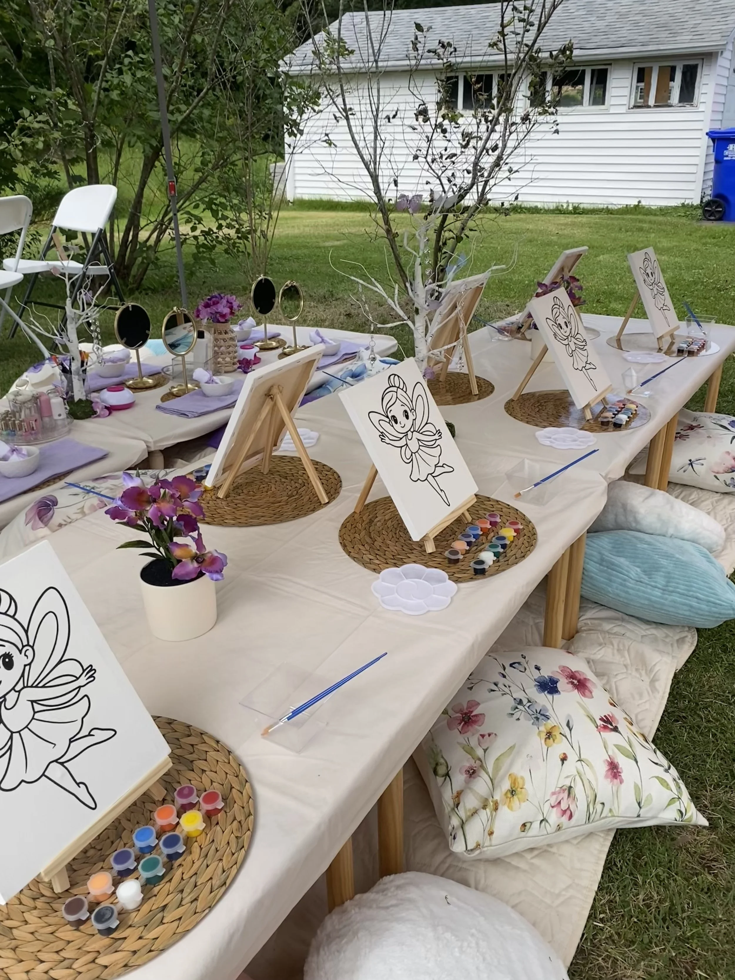 Outdoor craft setup with painting supplies, easels, and fairy-themed decorations on a table covered with a white cloth, with floral pillows and a small tree with fairy lights.