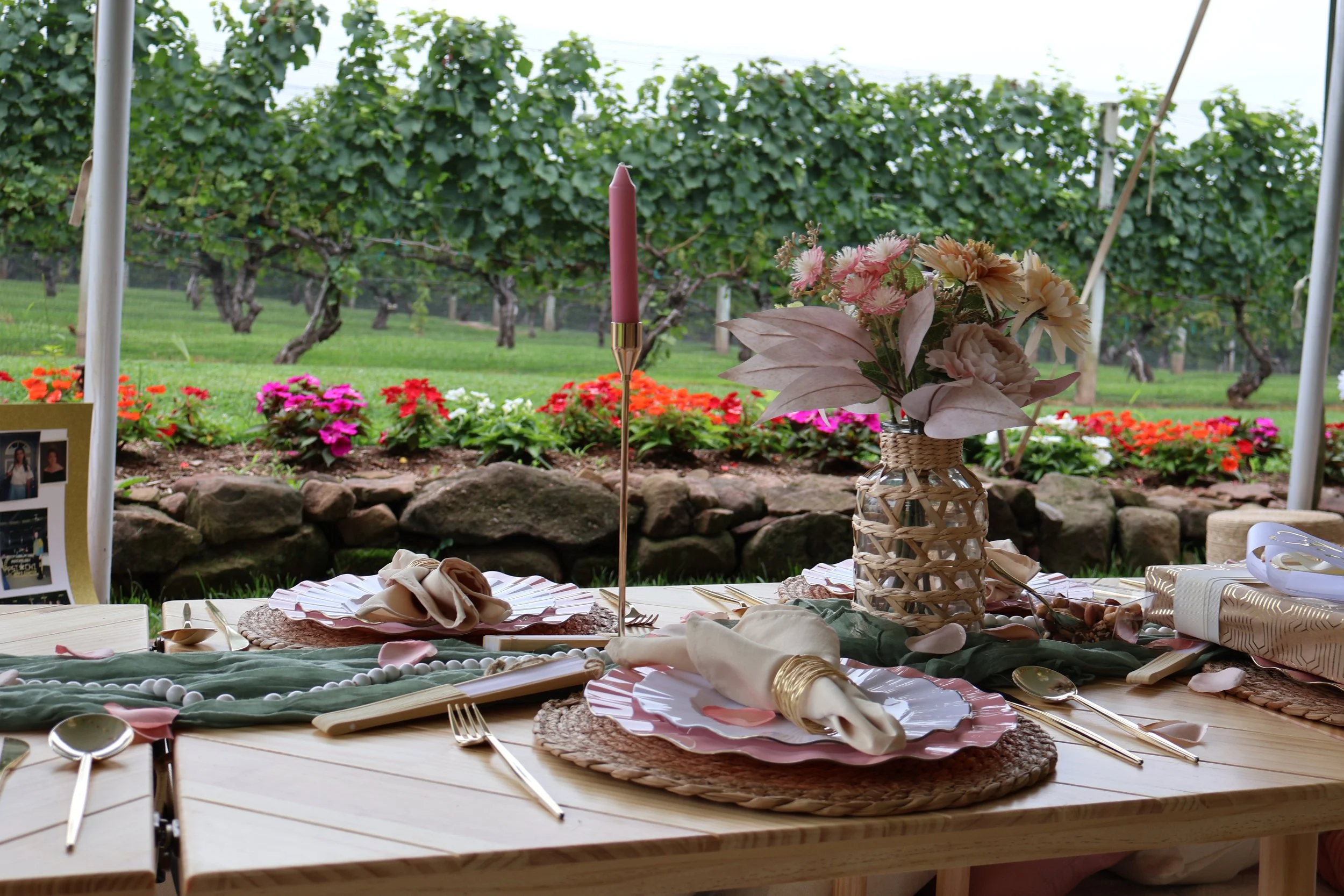 Table set for a gathering with floral centerpiece, plates, cutlery, napkins, and a pink candle in a decorative holder, overlooking a vineyard with rows of grapevines and colorful flowers.