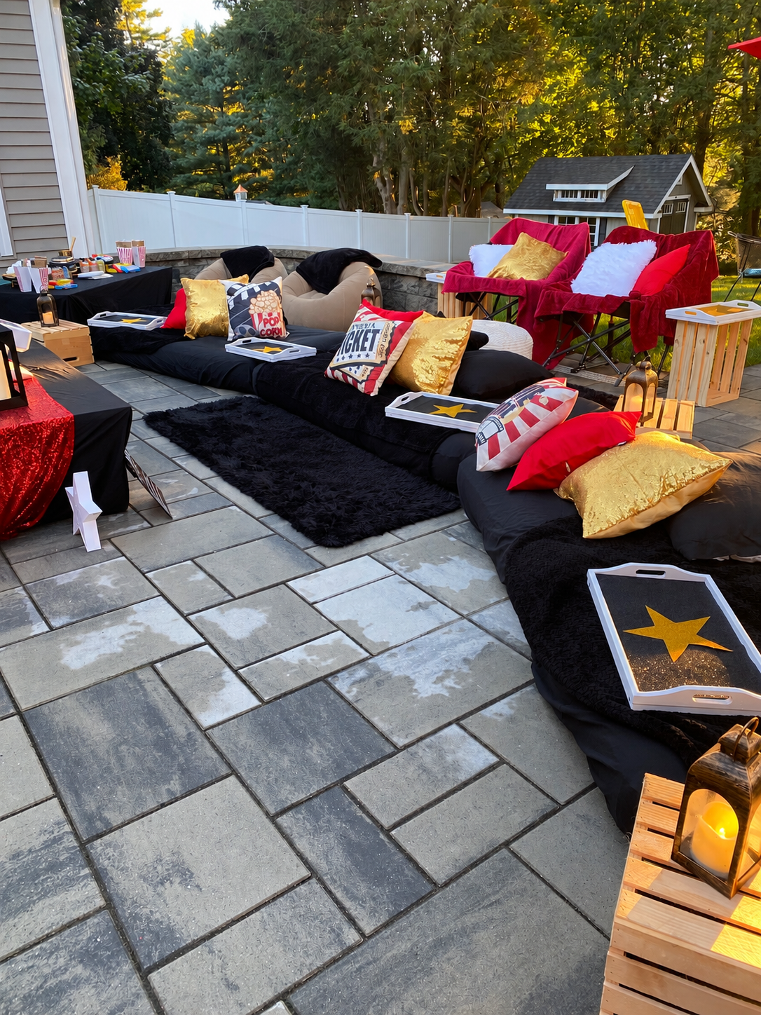 Outdoor backyard space decorated with red, white, black, and gold pillows, with a black blanket on the ground, and small tables containing lanterns and snacks, suggesting a party or gathering setup.