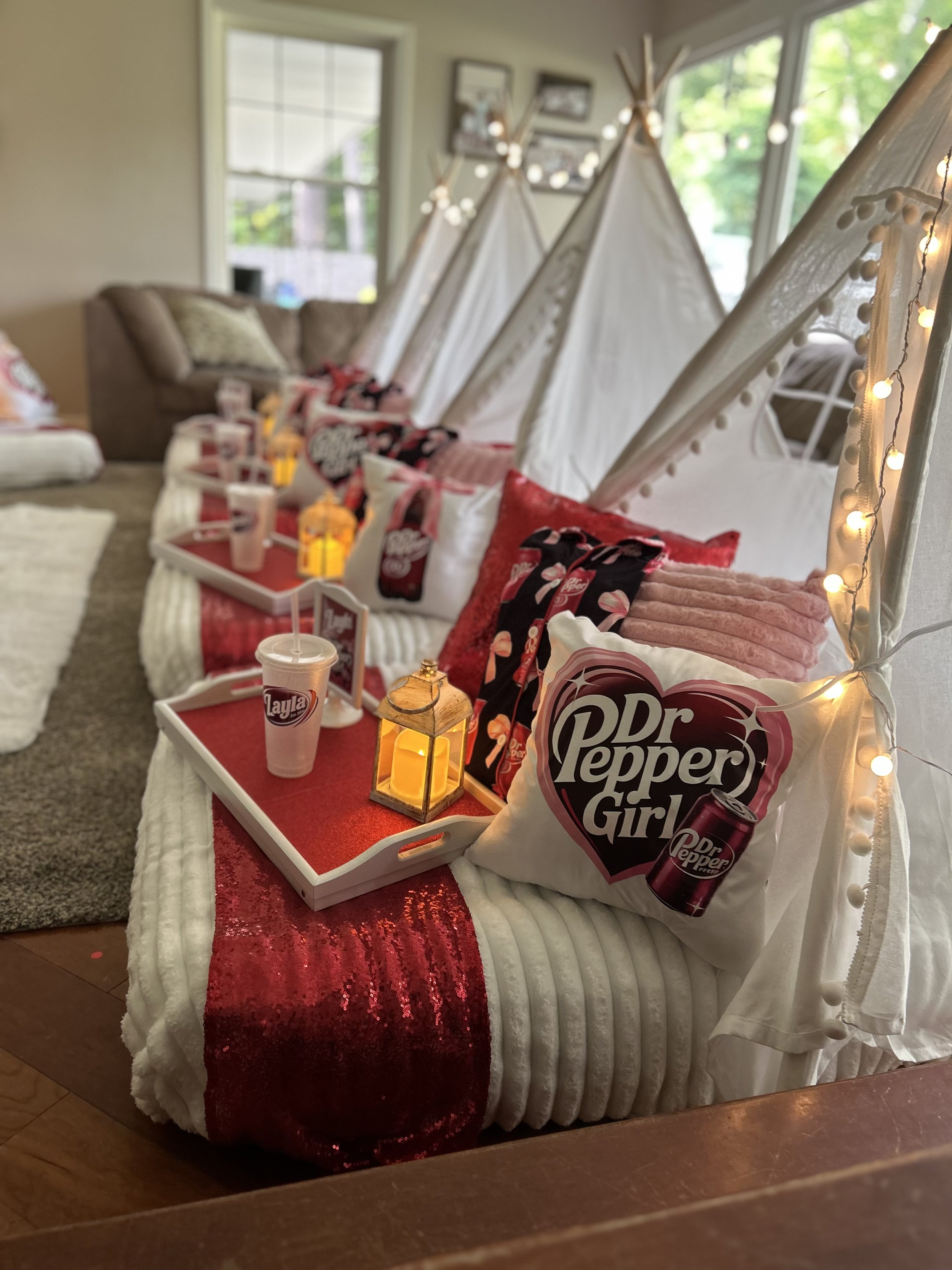 Party table with Dr Pepper Girl pillows, red sequin table runner, cups with straws, and decorative lanterns, set in a cozy living room with couches and large windows.