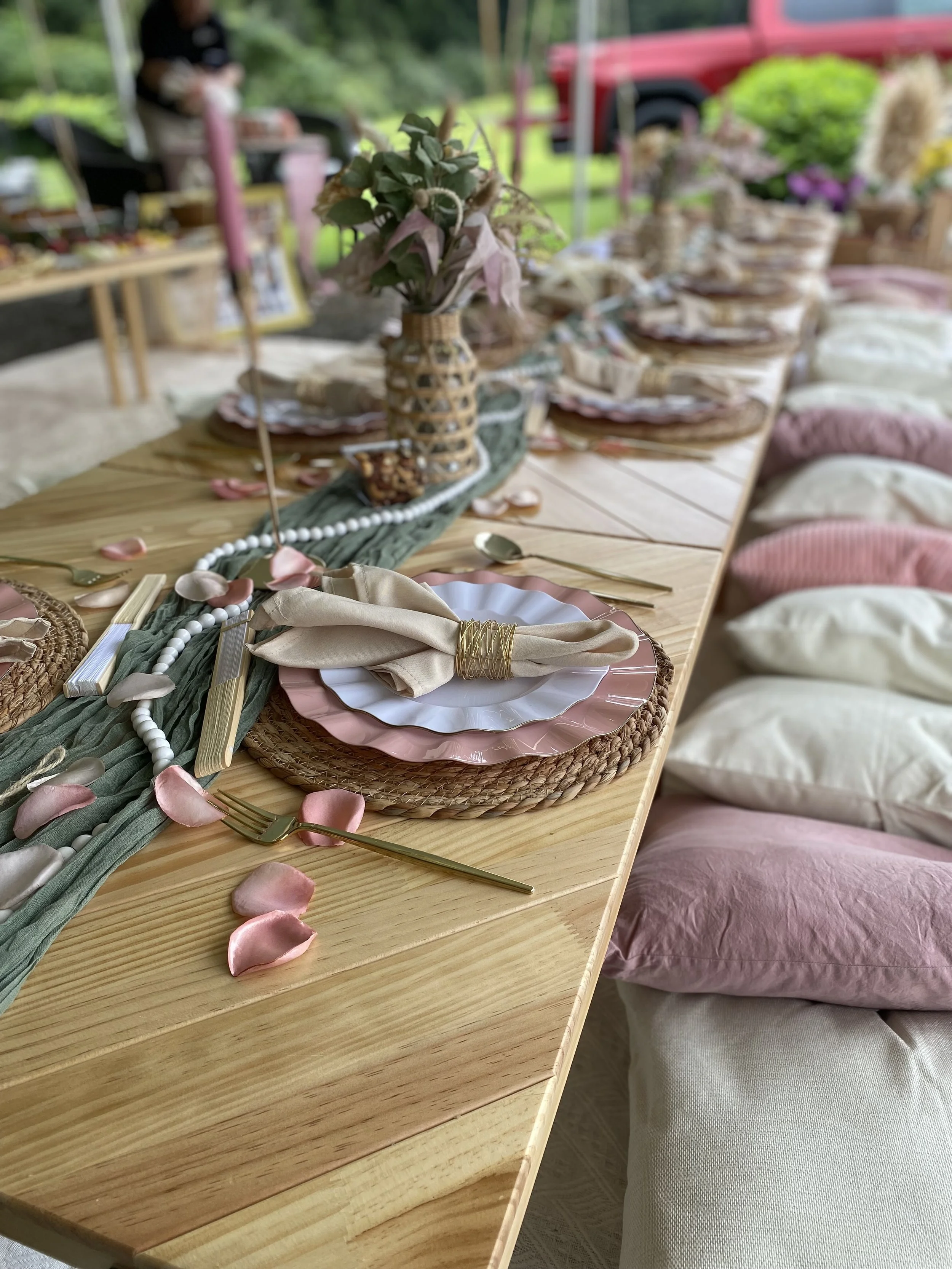 A wooden table set with pink and white plates, gold utensils, beige napkins, and pink rose petals. A vase with pink flowers and greenery decorates the center of the table, with a green table runner underneath. Cushions in pink and beige are placed on white chairs along the side of the table, set up outdoors.