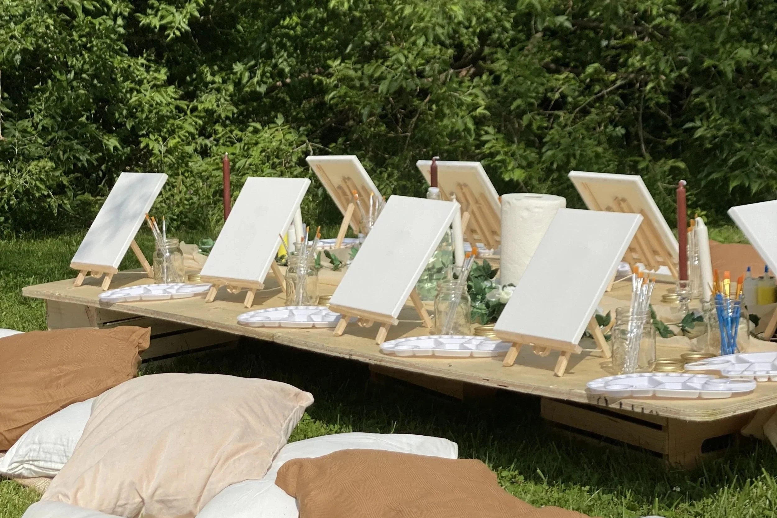 Set of small canvases on a long outdoor table, surrounded by paint supplies, water bottles, and cushions, with a green leafy background.