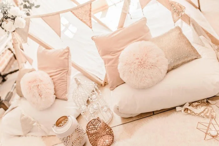 Cozy bed or seating area decorated with blush-colored pillows, fluffy round pillows, and stringed bunting under a tent-like canopy with lace and fabric accents, creating a feminine and elegant atmosphere.