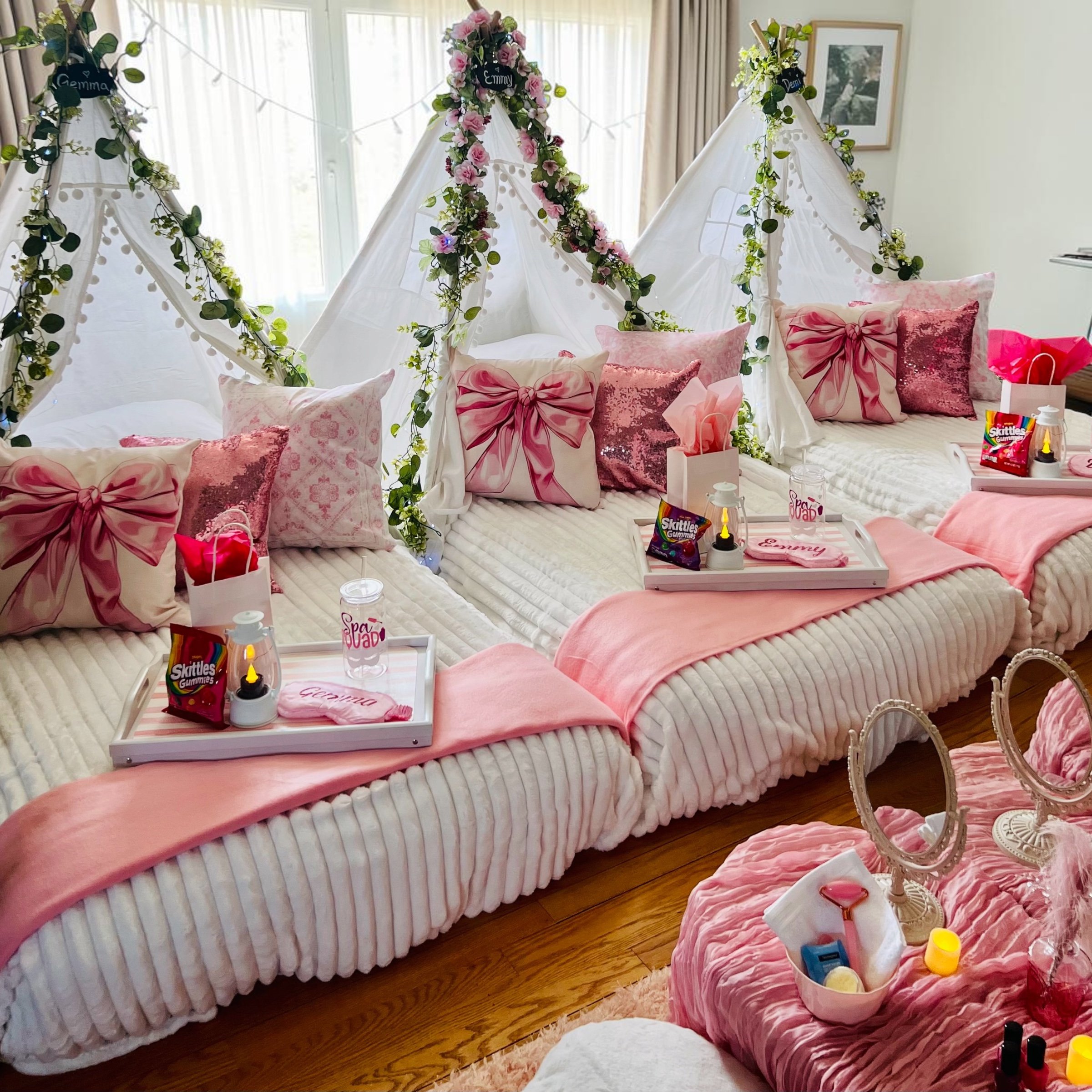Decorated beds with pink pillows and bows, floral garlands, gift bags, and party supplies for a celebration in a bedroom with large windows and framed artwork.