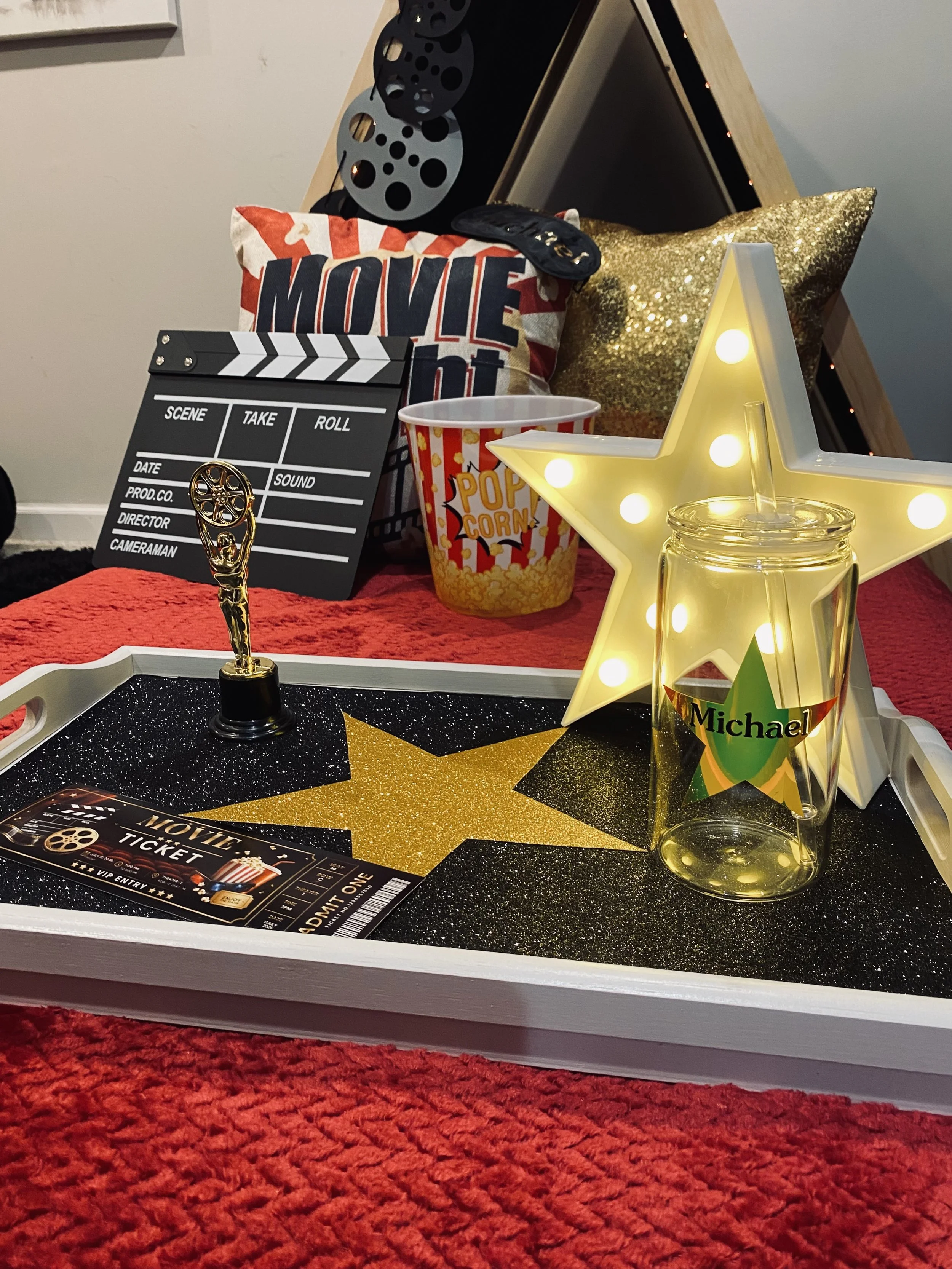 A table decorated with movie-themed items including a gold star-shaped light, a small trophy, a film slate, a popcorn container, movie tickets, and pillows, all placed on a red textured surface.