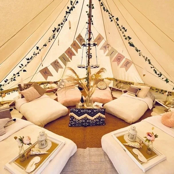 Interior of a large beige canvas tent with four beds and cozy decor, including pillows, lanterns, flowers, and a banner reading 'KINDNESS'.