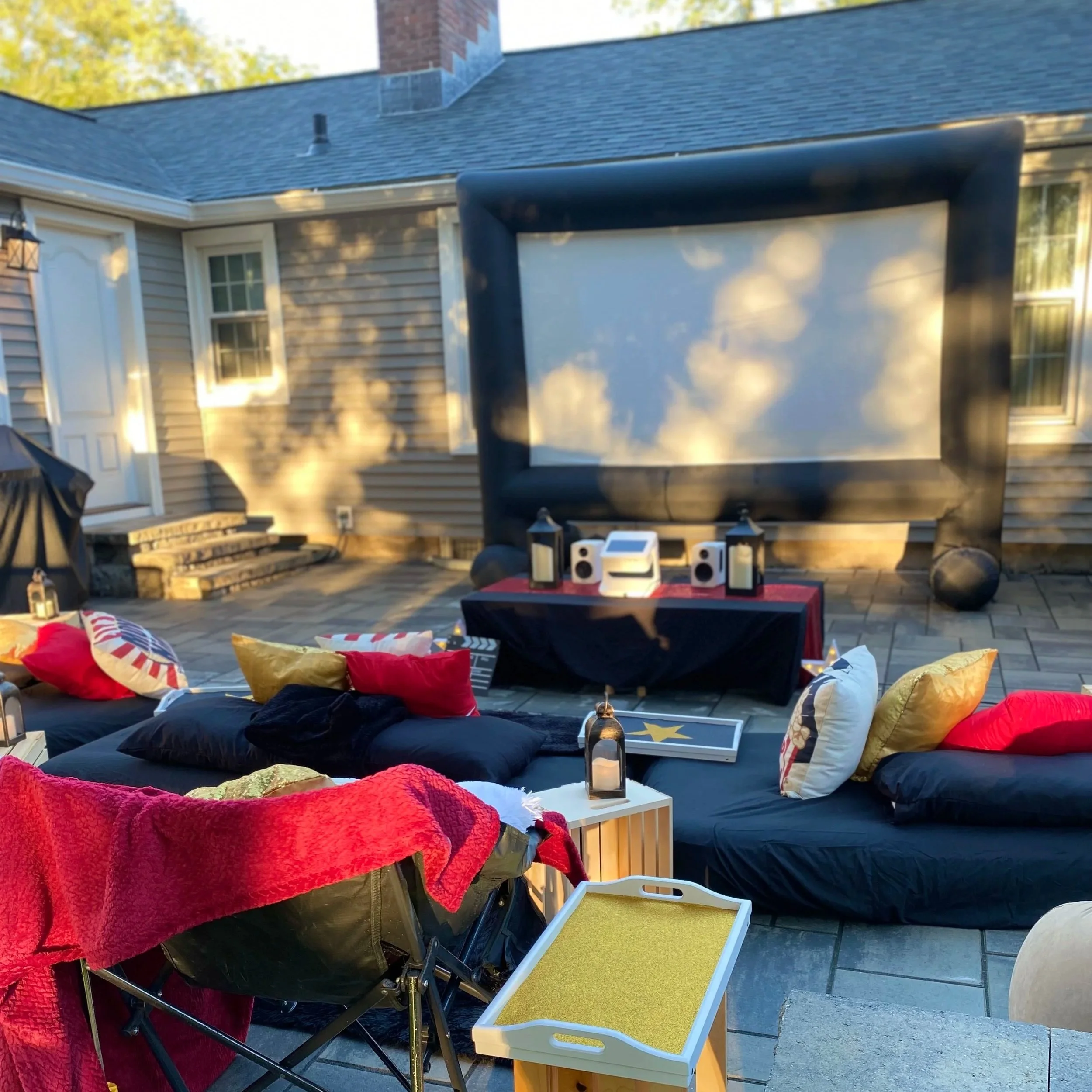 An outdoor backyard setting with a patio area, cushions, a small table with snack bags, a speaker, and a black trampoline. In the background, there's a house with grey siding and a blue shingled roof, and trees with green leaves.