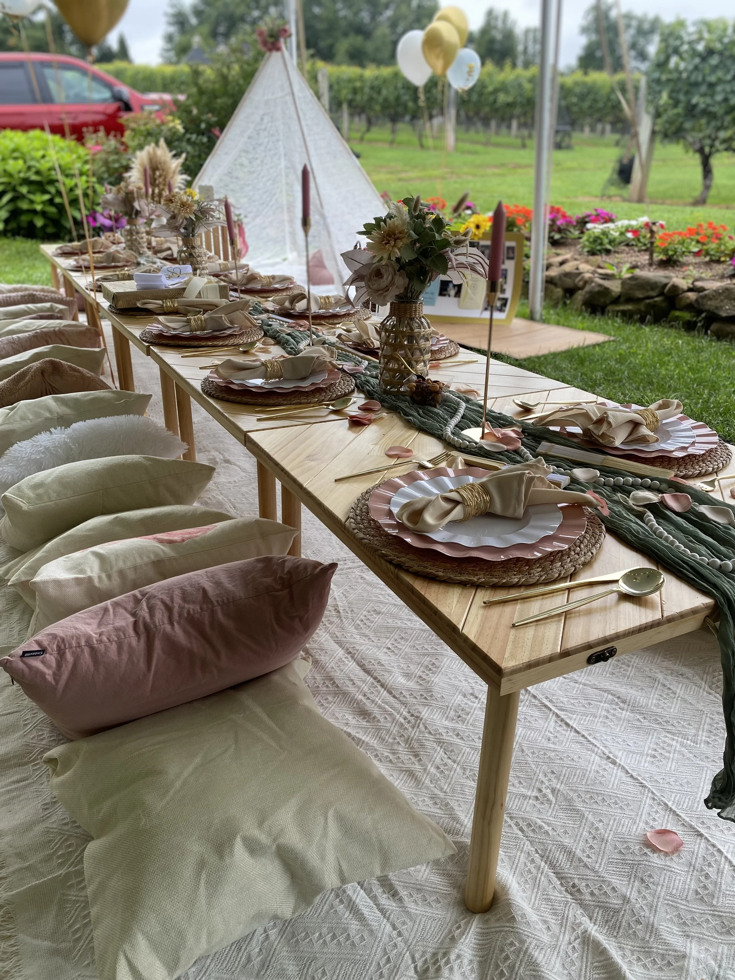 A long wooden table is set for a celebration outdoors, decorated with light pink and cream napkins, gold cutlery, pink candles, and centerpieces with flowers and pearl strings. Behind the table, there is a white teepee tent with pink flowers, and bal