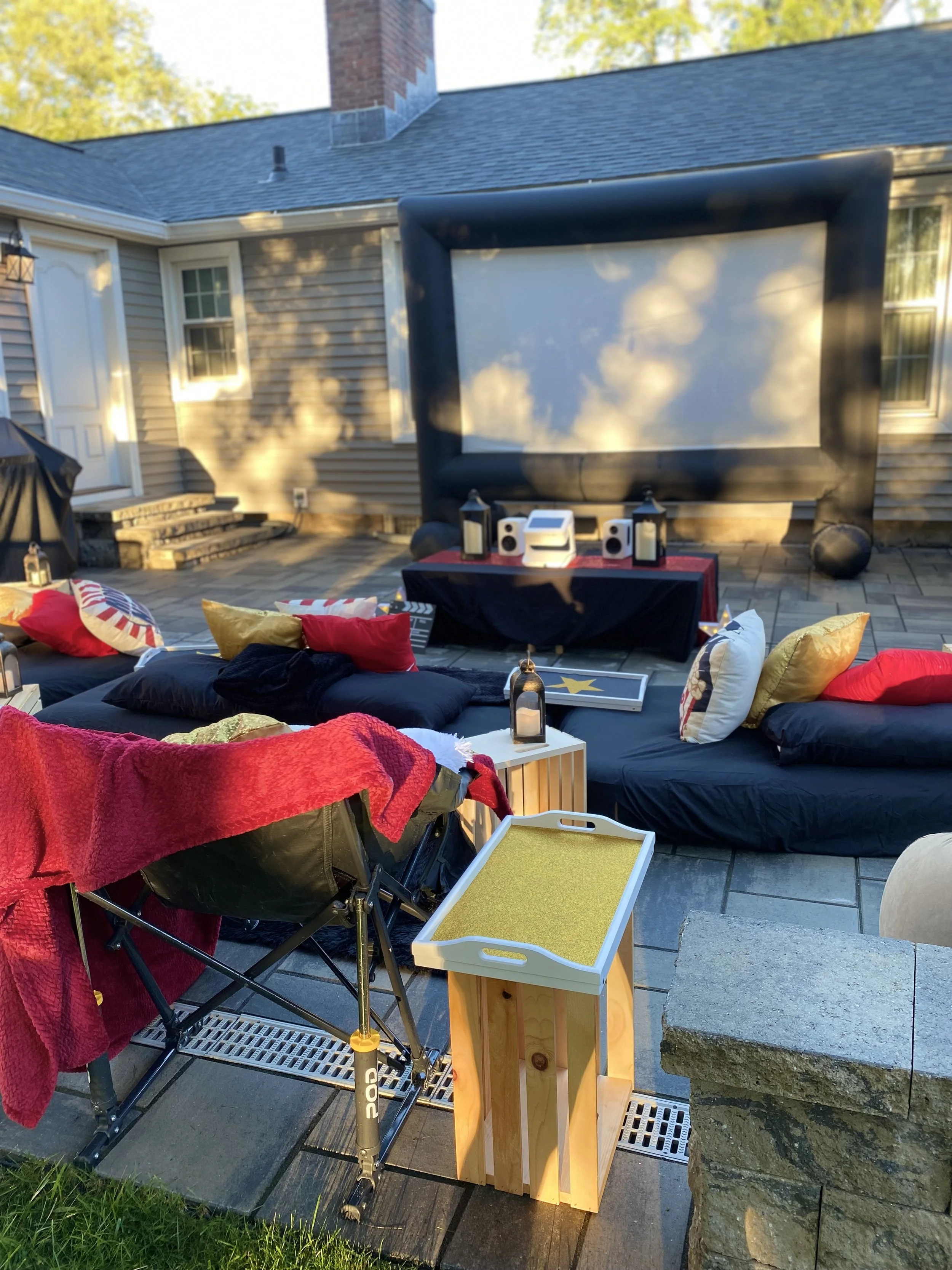 Outdoor backyard with various chairs, pillows, and a large inflatable outdoor movie screen set up for an outdoor movie night. There are lanterns and a small wooden side table among the seating area.