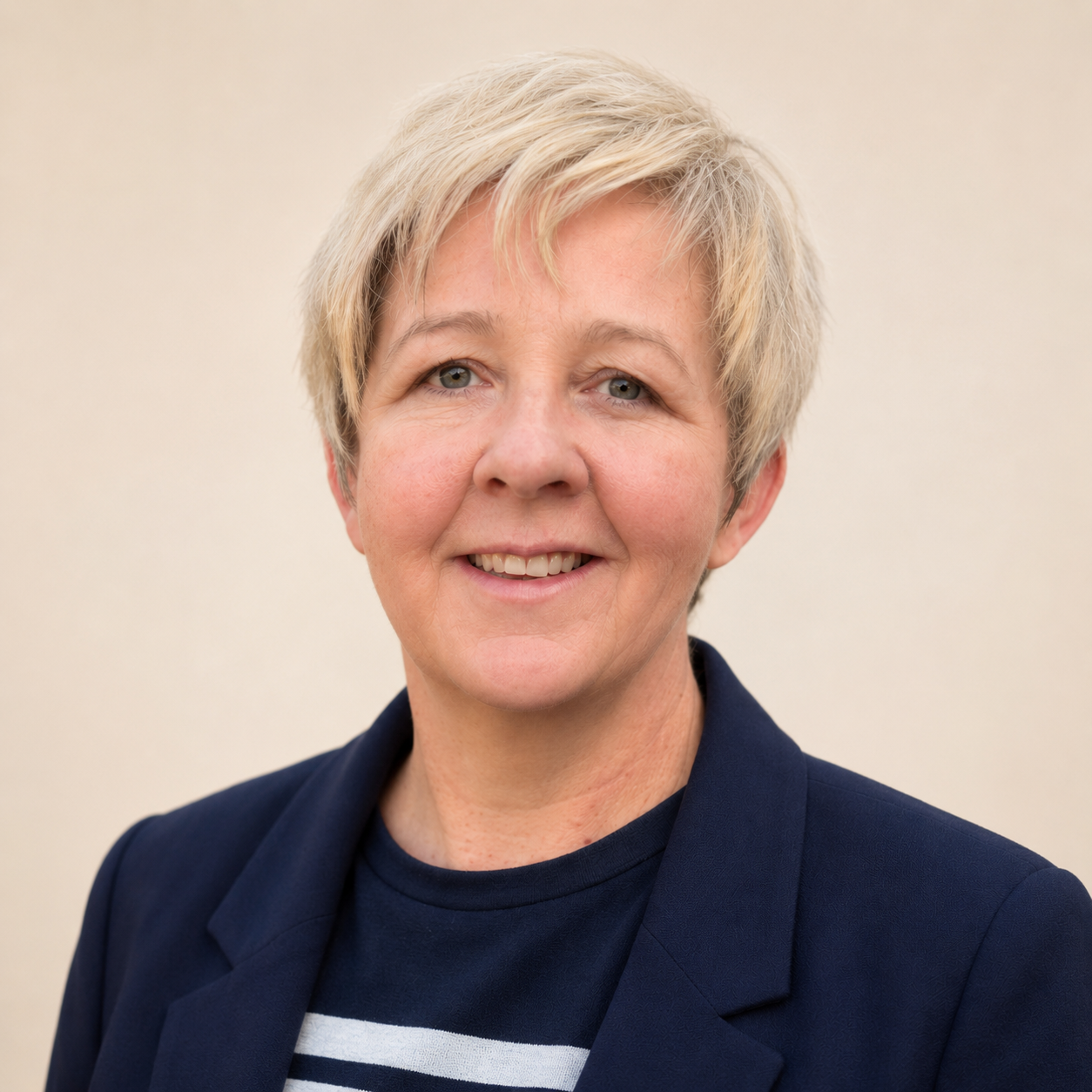 A smiling middle-aged woman with short blonde hair wearing a navy blazer and a striped shirt against a plain light background.