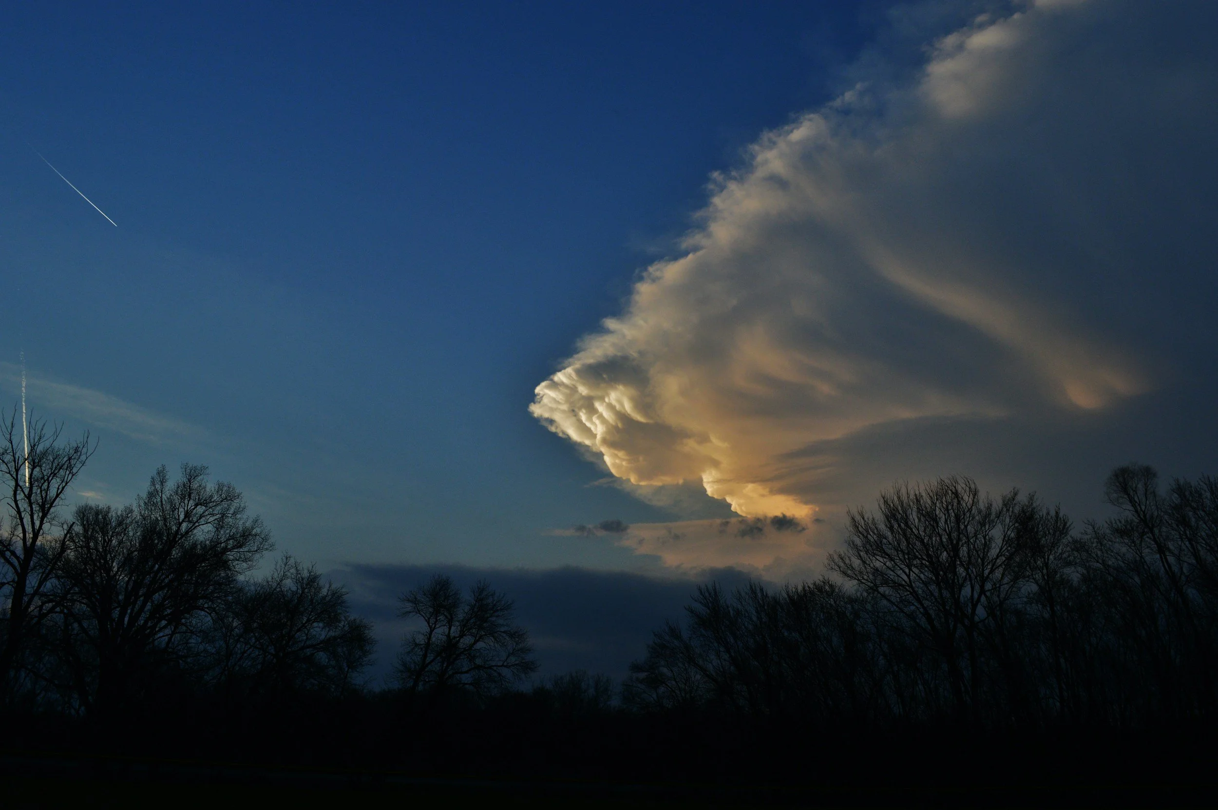 Der Himmel ist teilweise bedeckt mit großen, dunklen Wolken, und die Sonne scheint durch die Wolken. Es sind Laubbäume ohne Blätter im Vordergrund zu sehen.