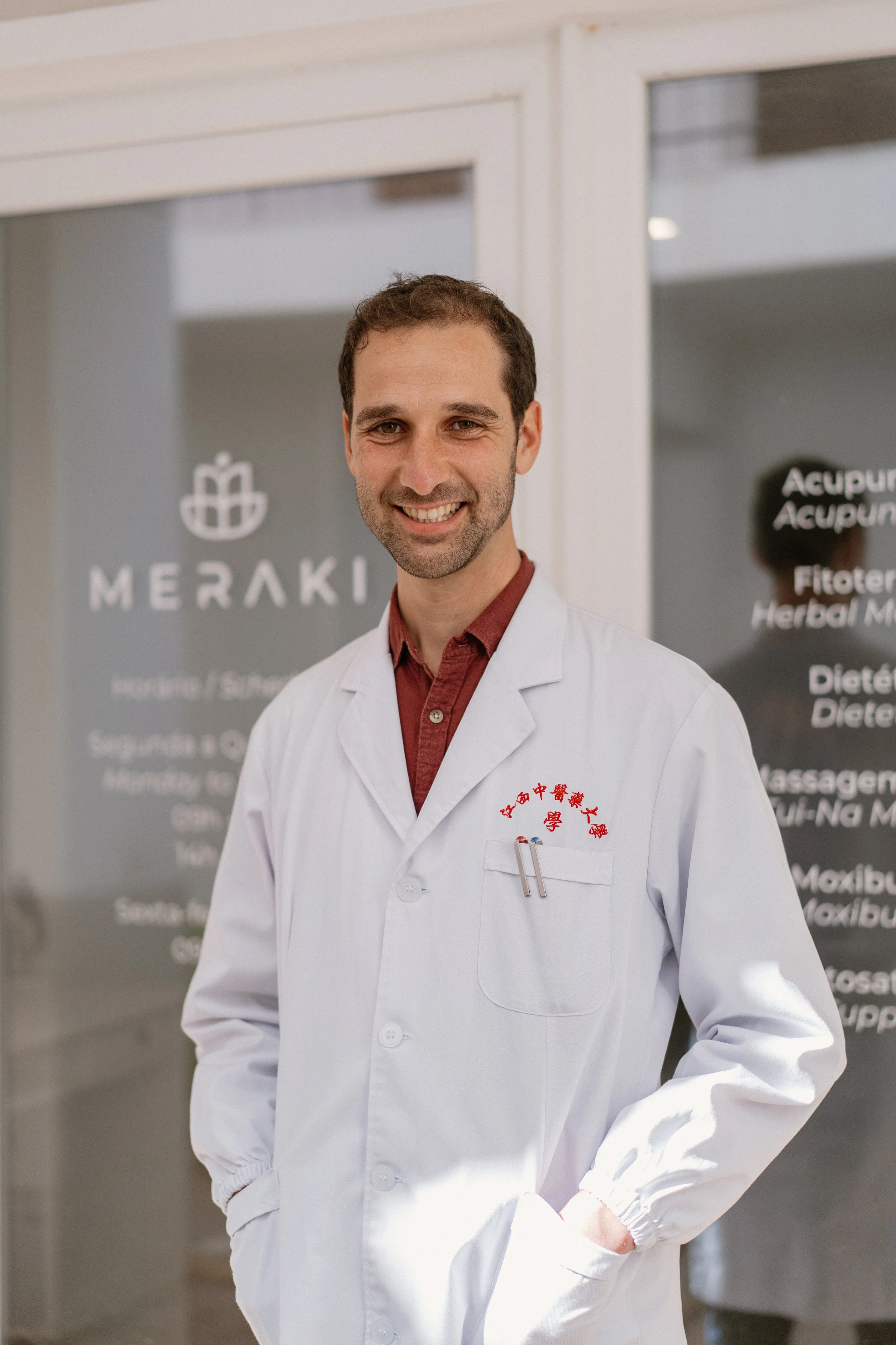 A man wearing a white lab coat with red Chinese characters, standing in front of a glass door with logo and text for Meraki Holistic, smiling at the camera.