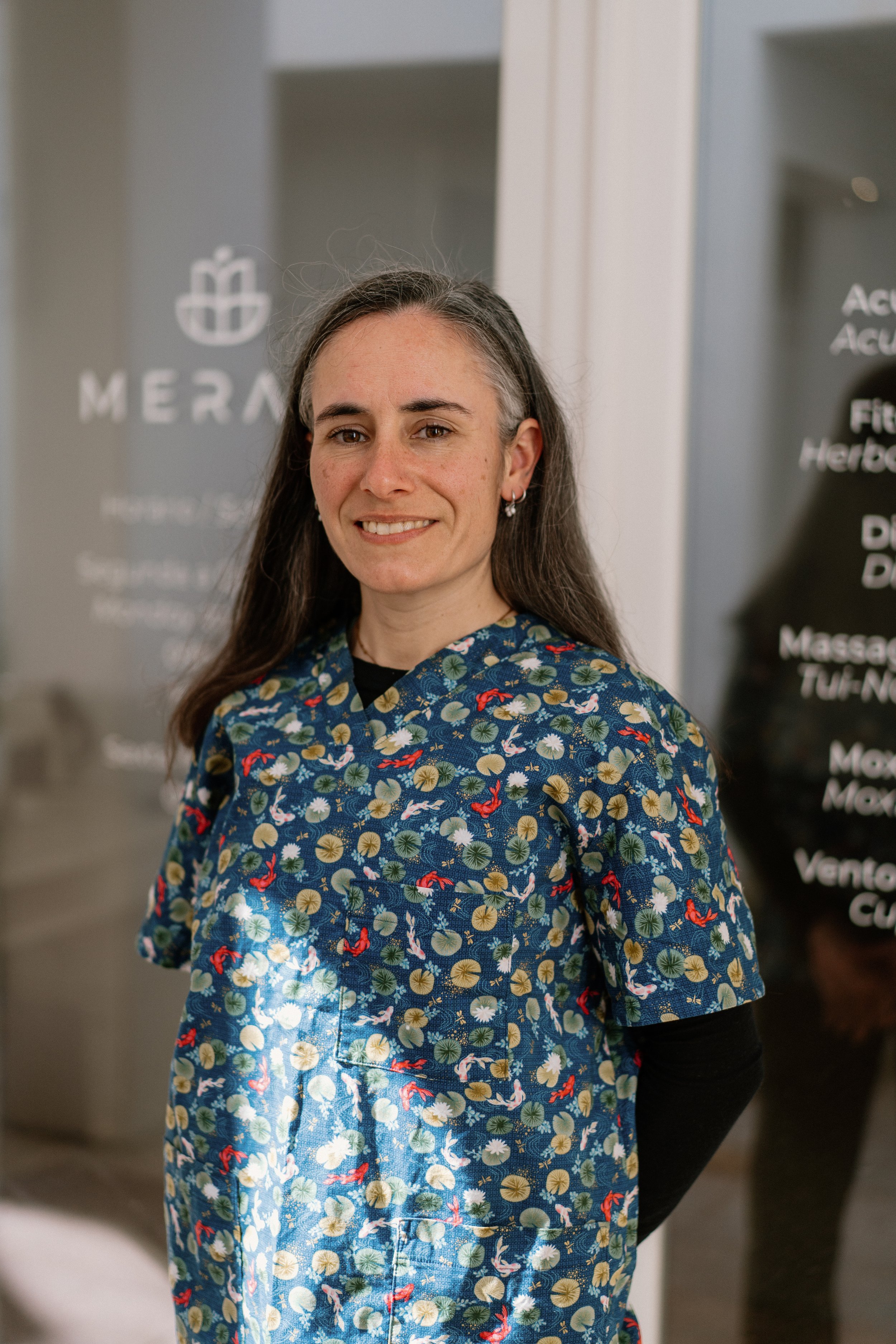 A woman with long gray hair wearing a colorful, patterned scrub top standing indoors. In the background, blurred signage features the word 'MERAK' and an icon of a beaker or flask.